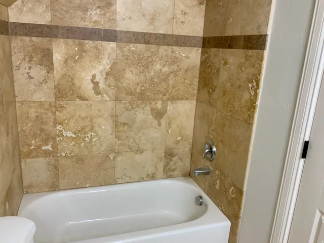 White bathtub with chrome fixtures, surrounded by light-toned natural stone-look wall tiling