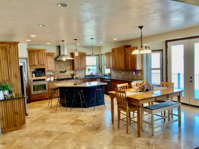 Spacious kitchen featuring stone tile flooring, warm wood cabinetry, a dark-toned island with integrated seating, stainless steel appliances, and double glass doors leading to an outdoor space