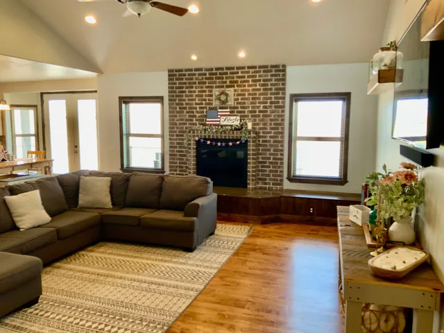 Spacious living area featuring a vaulted ceiling with recessed lighting and a ceiling fan, a brick-surround fireplace with a hearth, wood-finish flooring, and natural light from multiple windows