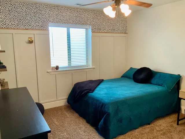 Bedroom featuring board and batten wall paneling, a window with horizontal blinds, textured carpeting, and a ceiling fan with light fixture