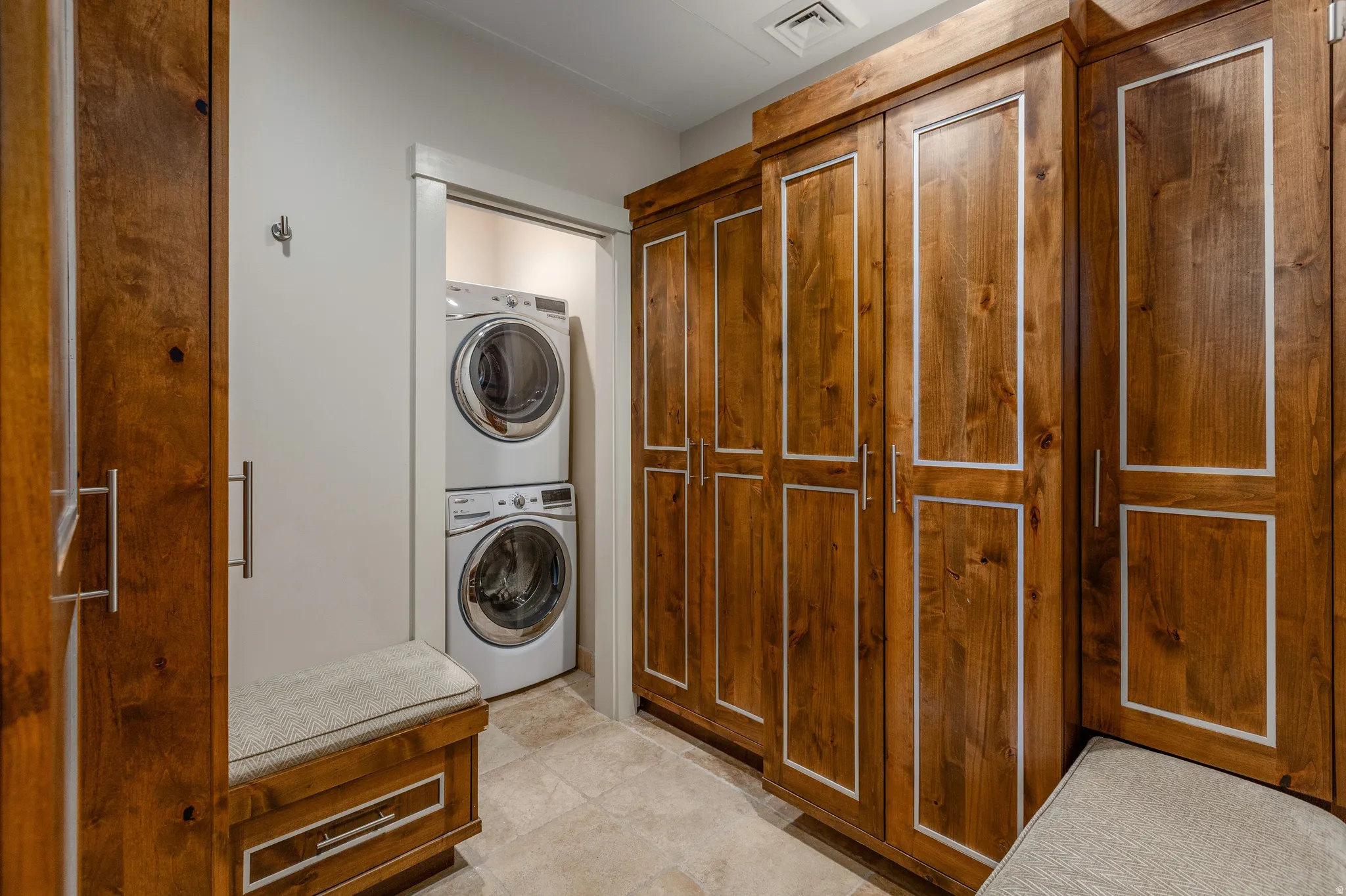 Dedicated laundry area featuring a stacked washer and dryer, extensive wood-finish cabinetry with contrasting trim, integrated bench seating with upholstered cushions, and tile flooring