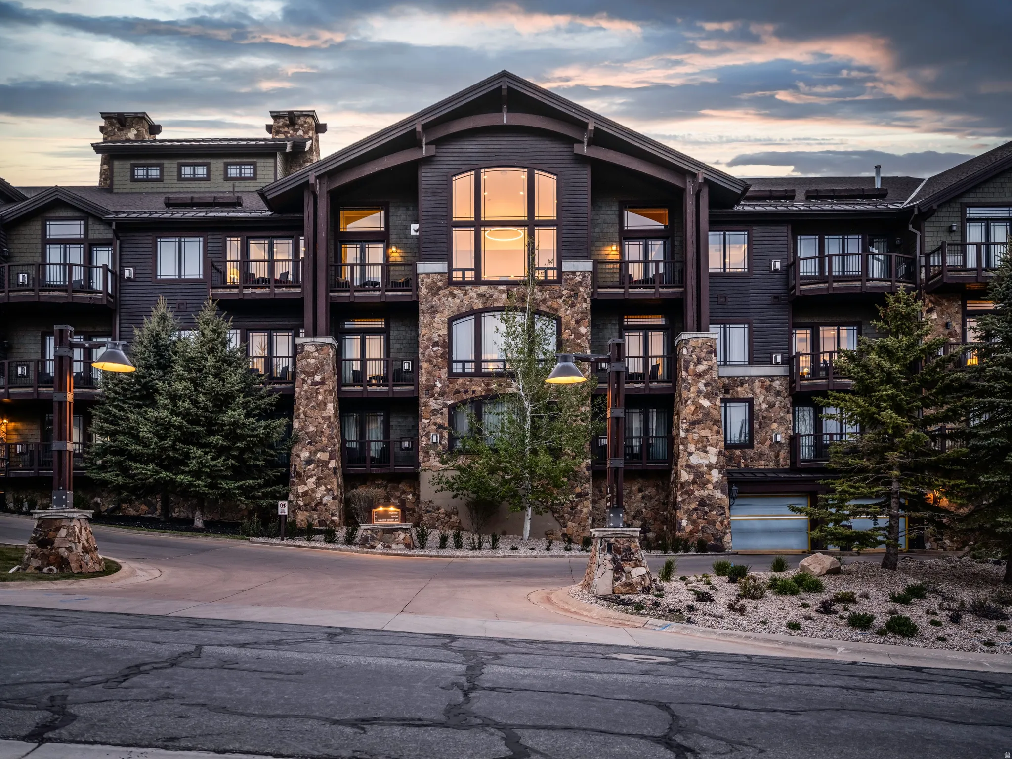 Multi-level structure featuring extensive stone and wood siding, multiple balconies, a grand central window arrangement, integrated garage access, and mature evergreen landscaping