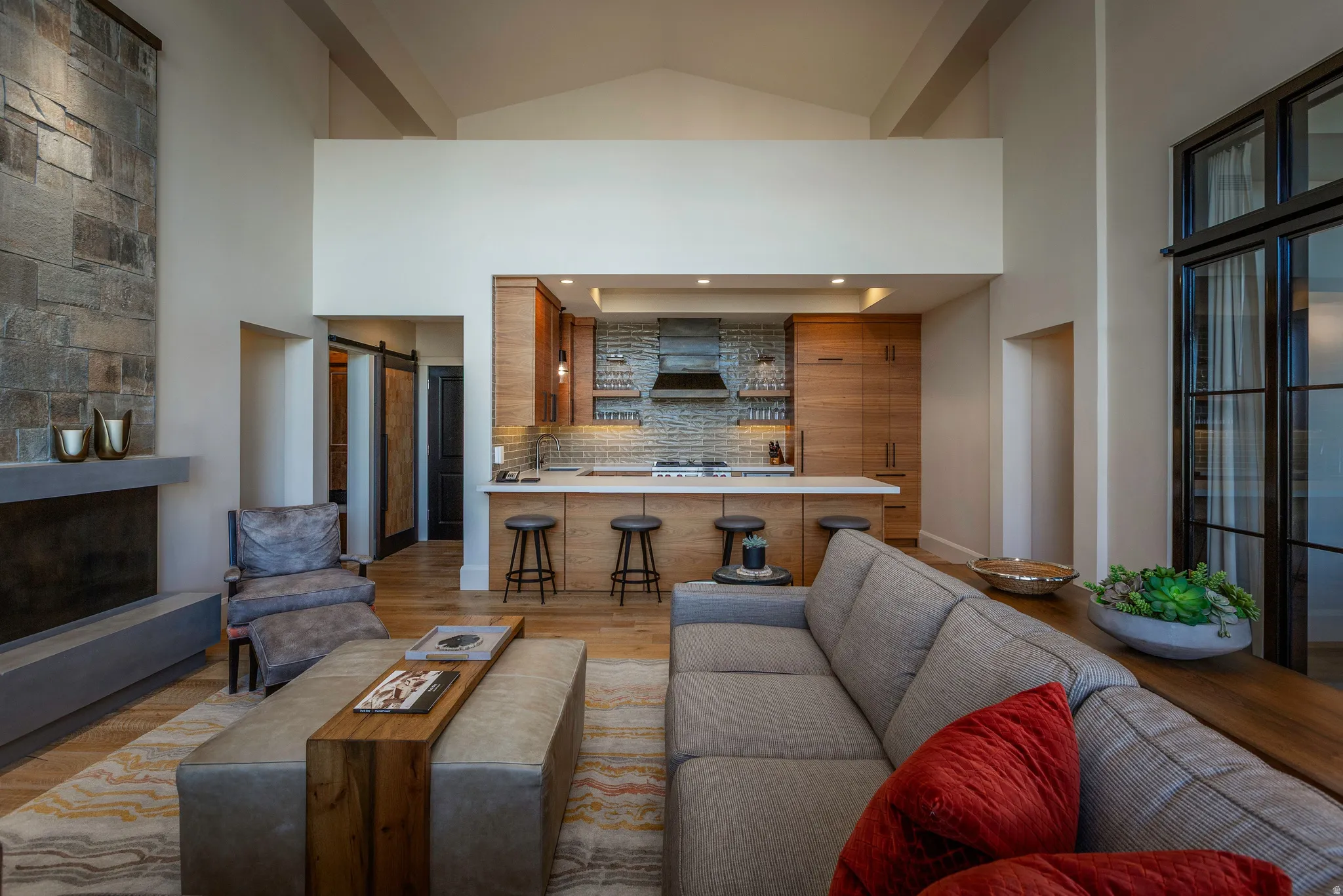 Open-concept living space featuring a floor-to-ceiling stone fireplace, a kitchen with wood cabinetry and a white countertop, and high vaulted ceilings with exposed beams
