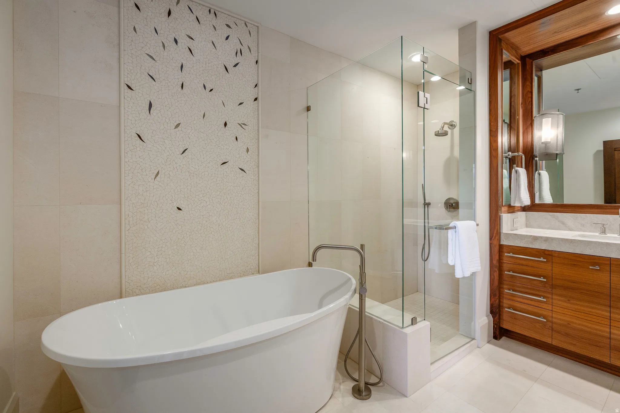 Freestanding soaking tub with floor-mounted faucet, glass-enclosed shower, wood-finish vanity with stone countertop, large mirror, and tiled accent wall