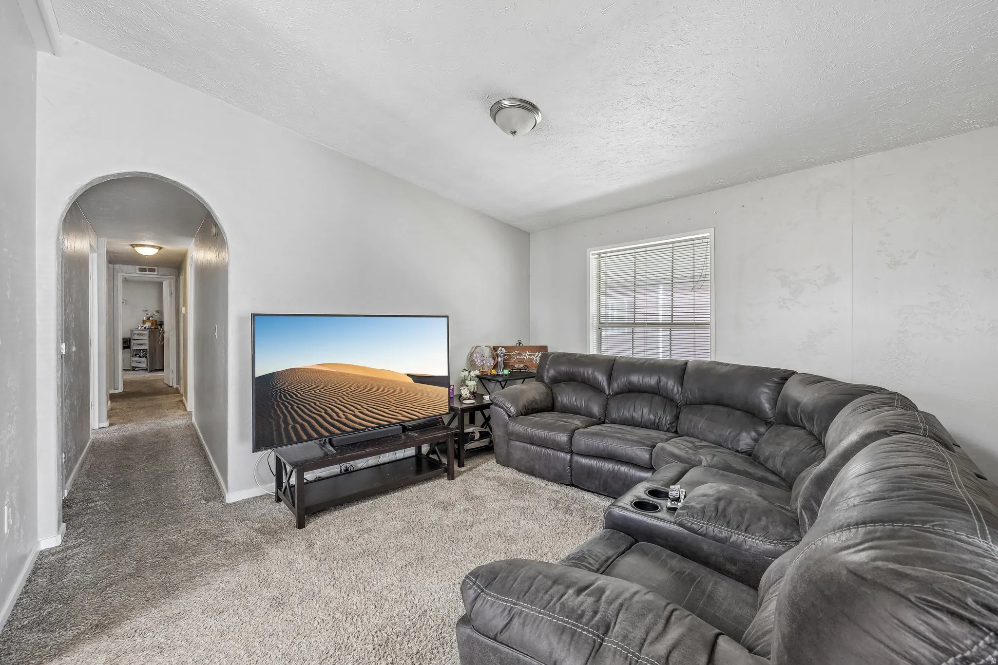 Carpeted living area featuring a large window with blinds, a prominent arched doorway, and light-toned walls