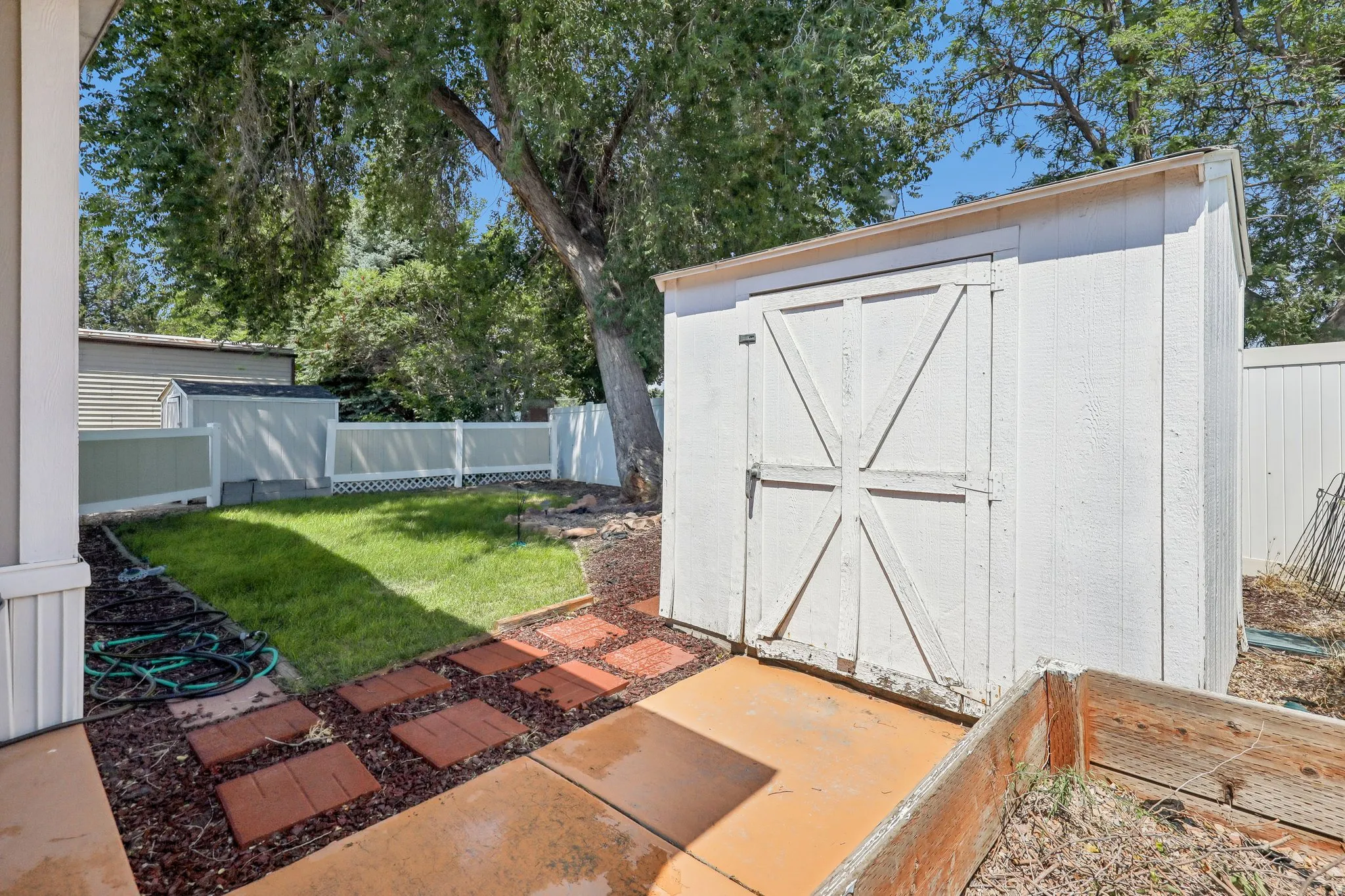 White storage shed with a panel door, concrete patio, and brick paver pathway