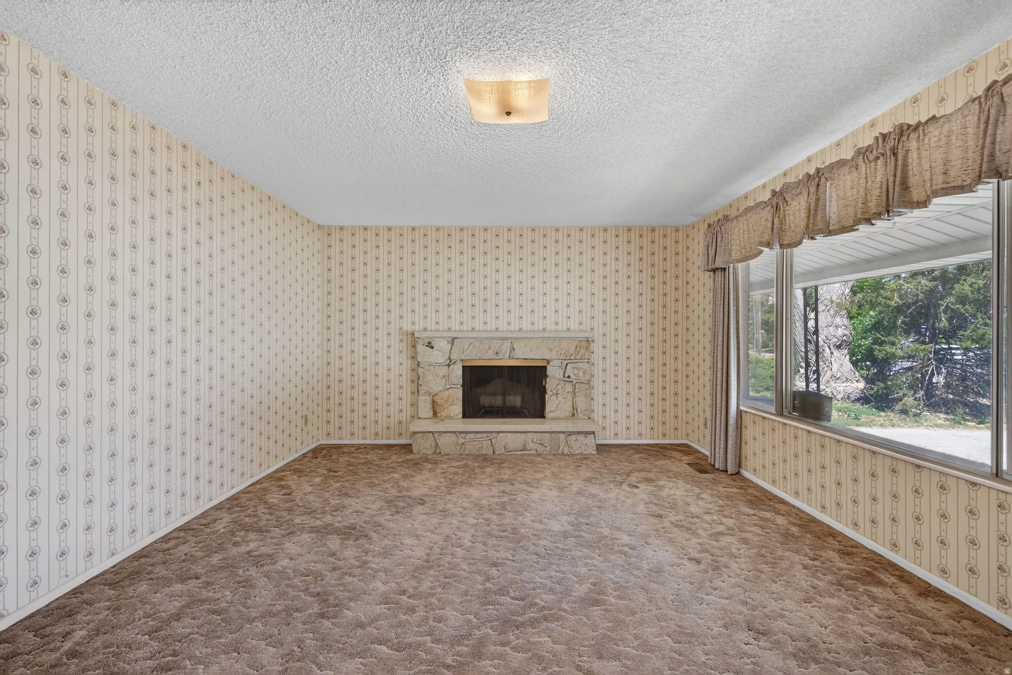 Spacious living area featuring a stone fireplace, patterned wallpaper, textured ceiling, and a large bay window