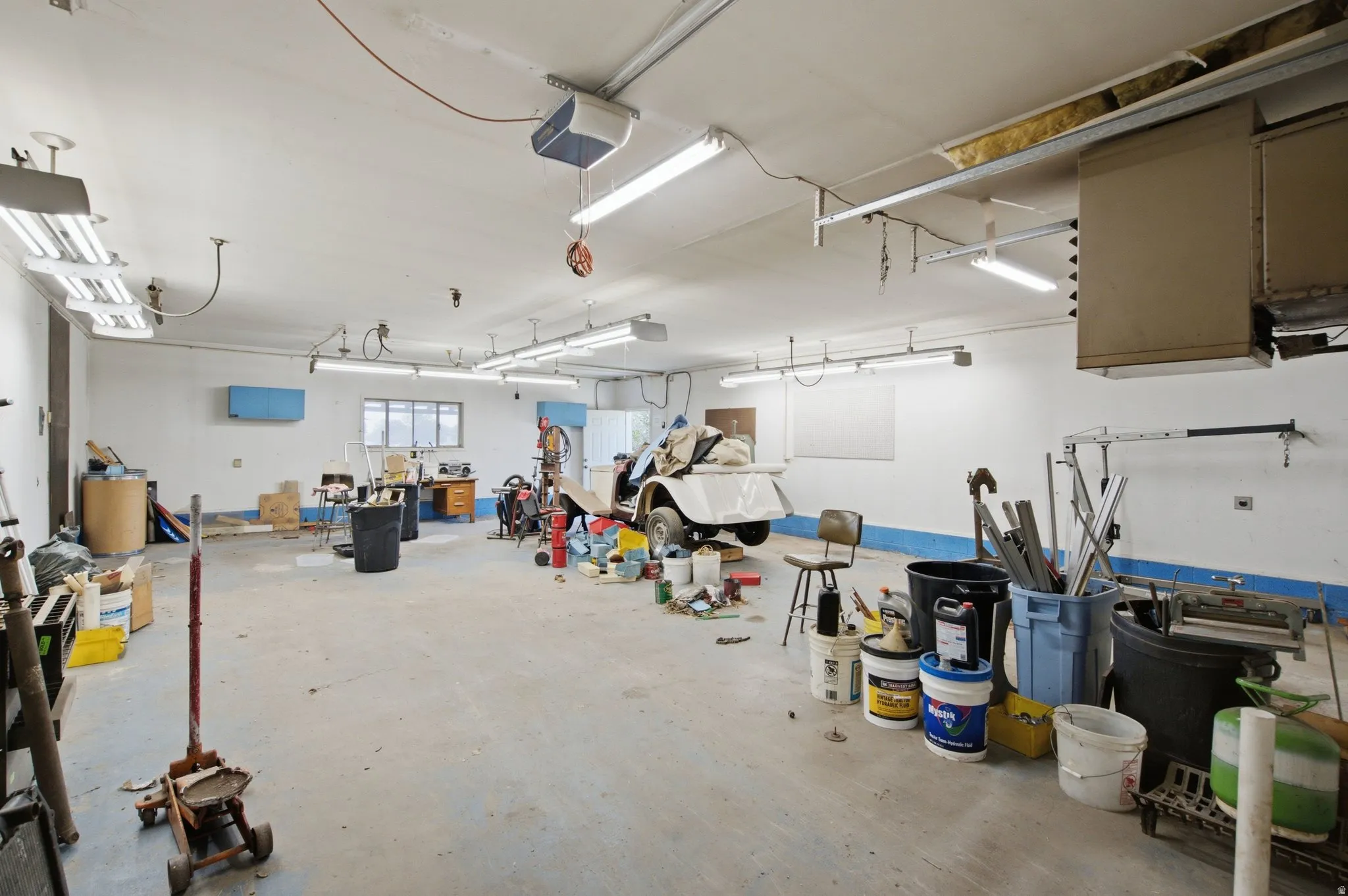Spacious garage with concrete flooring, multiple fluorescent lighting fixtures, exposed overhead ductwork, and window openings
