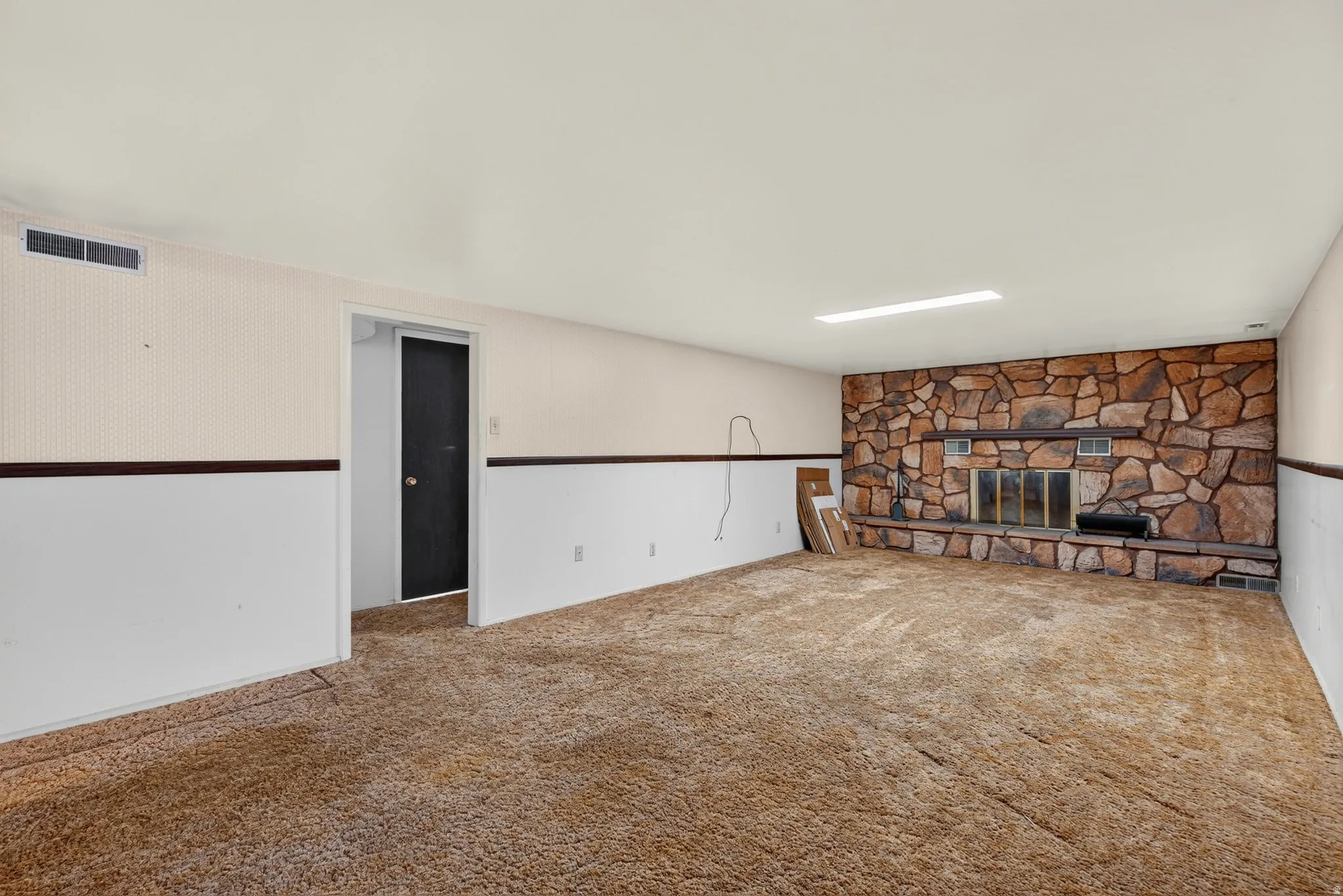 Spacious room featuring a full-wall stone fireplace with hearth, wall-to-wall carpeting, a dark interior door, and wainscoting with patterned wallpaper above