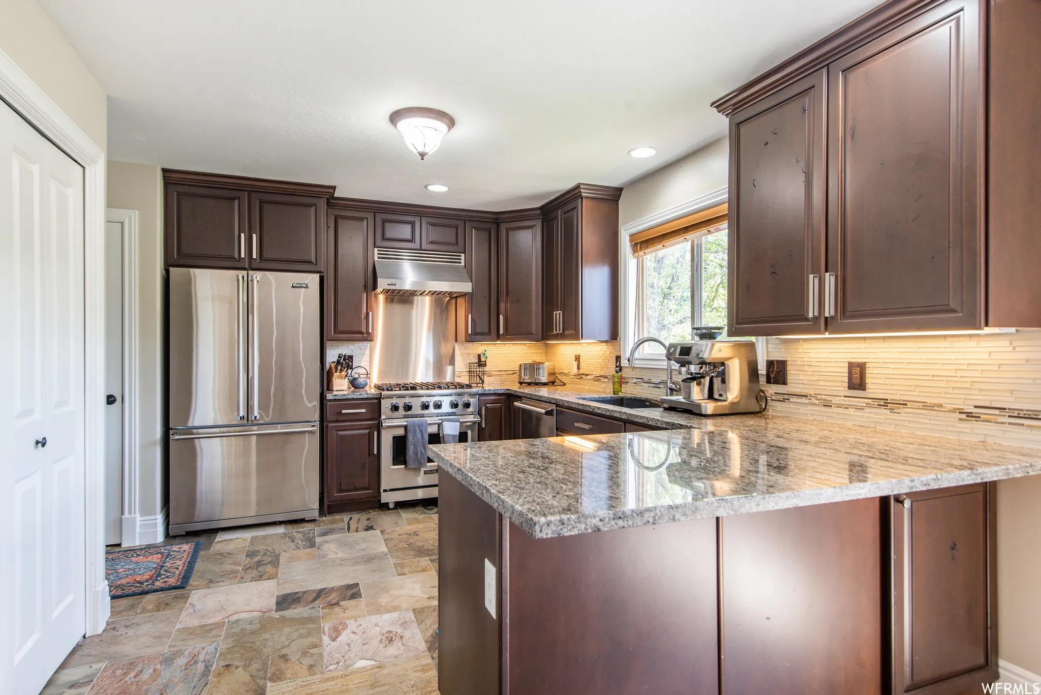 Kitchen featuring dark wood cabinetry, stainless steel appliances, speckled stone countertops, a tile backsplash, and multi-toned stone tile flooring