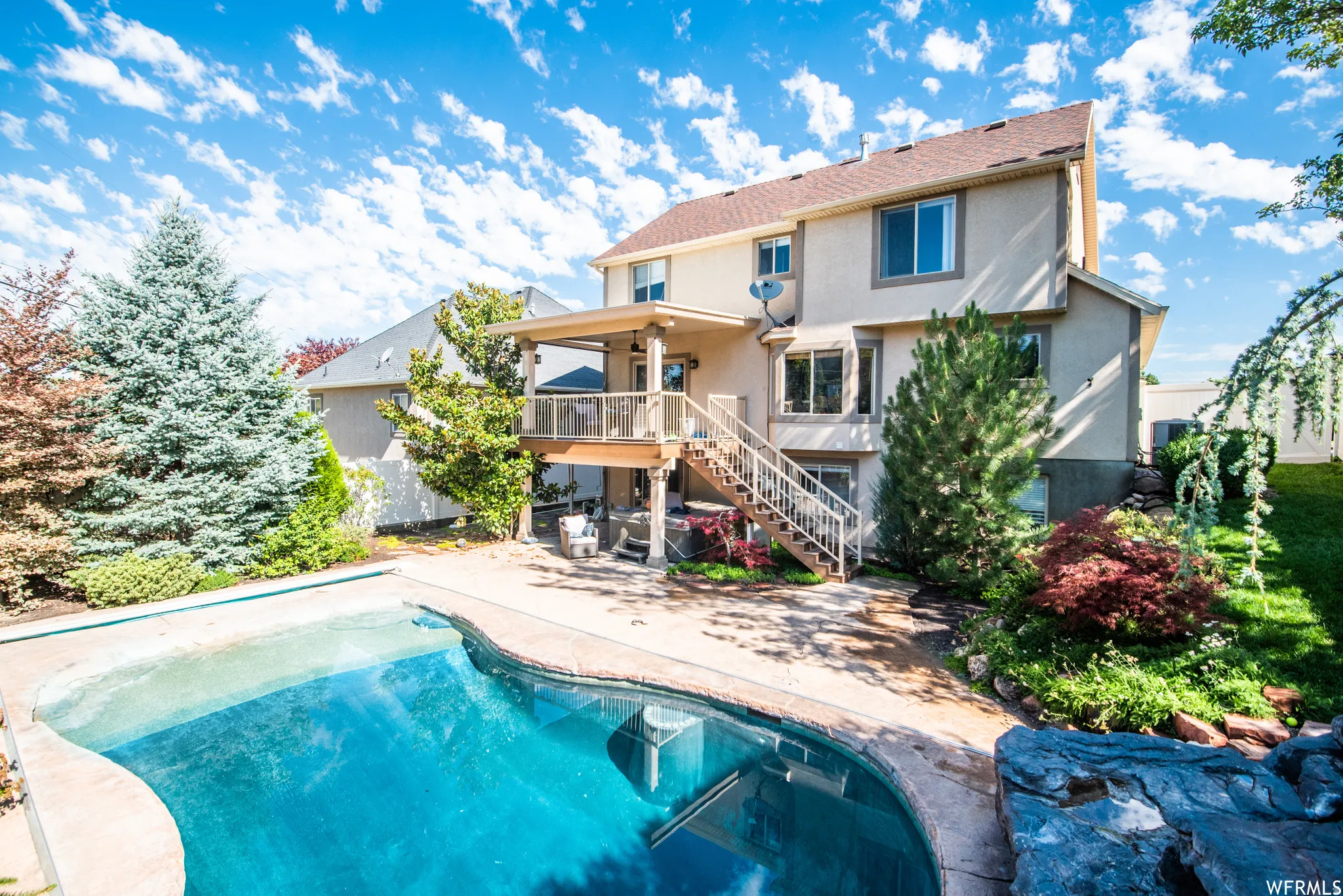 Rear exterior featuring an irregular-shaped in-ground pool with concrete surround, two-story deck with covered patio, and mature landscaping