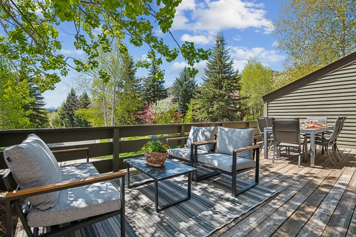 Spacious outdoor deck featuring wood flooring, a wooden railing, and a slatted exterior wall, surrounded by mature trees