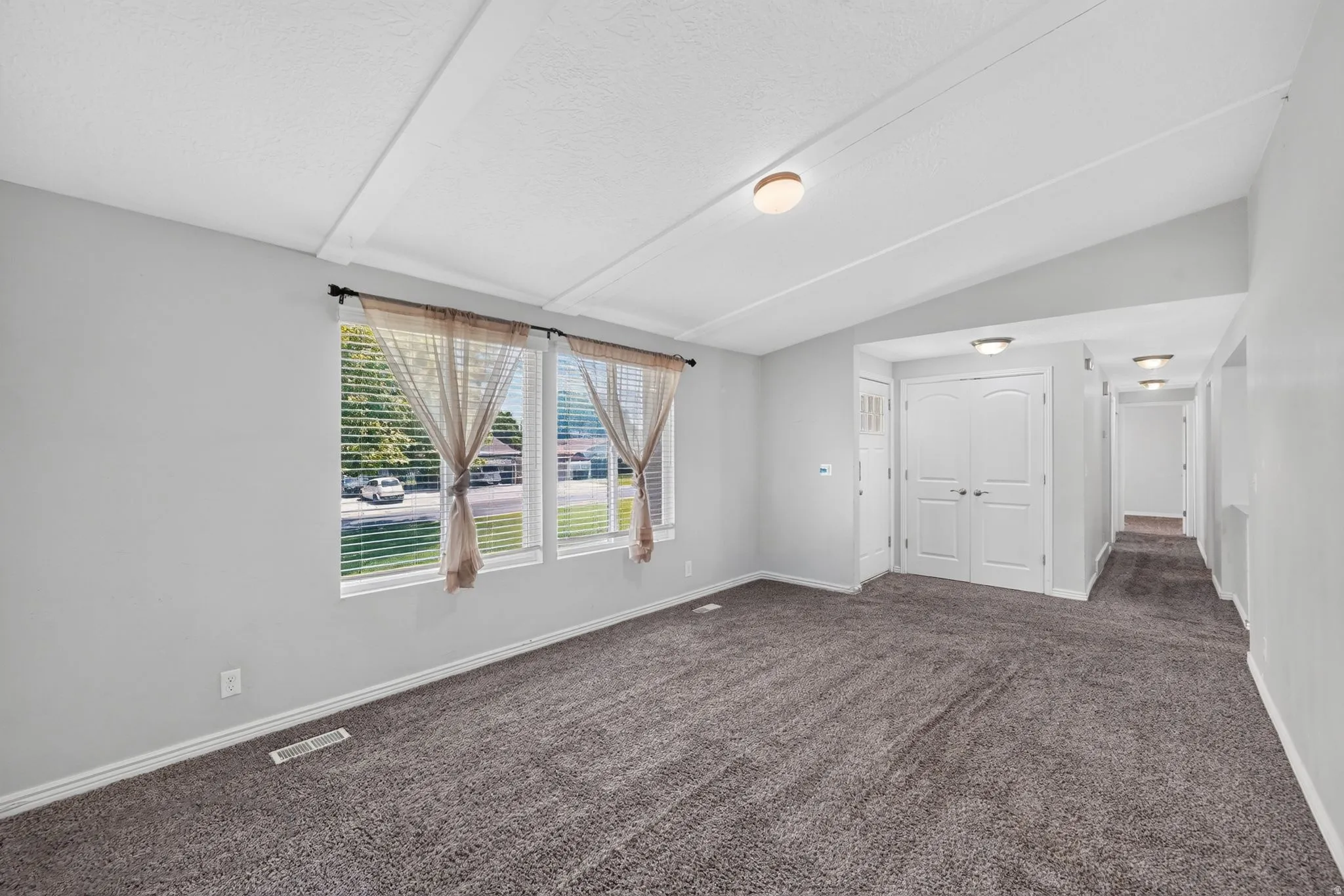 Open-concept living area featuring a vaulted ceiling with exposed beams, soft gray wall paint, and charcoal-toned carpeting