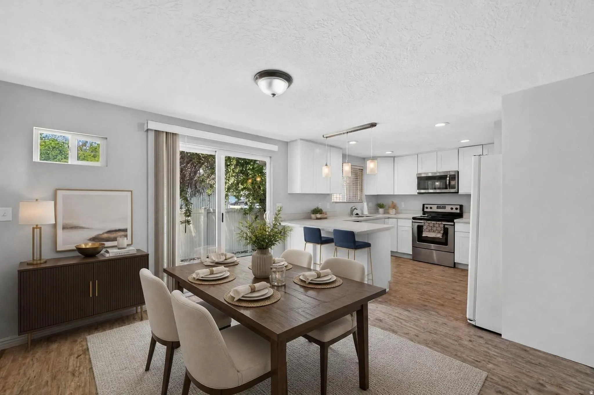 Open-concept living area with wood-finish flooring, a contemporary dining table, sliding glass door access to the exterior, and a kitchen featuring white cabinetry and stainless steel appliances