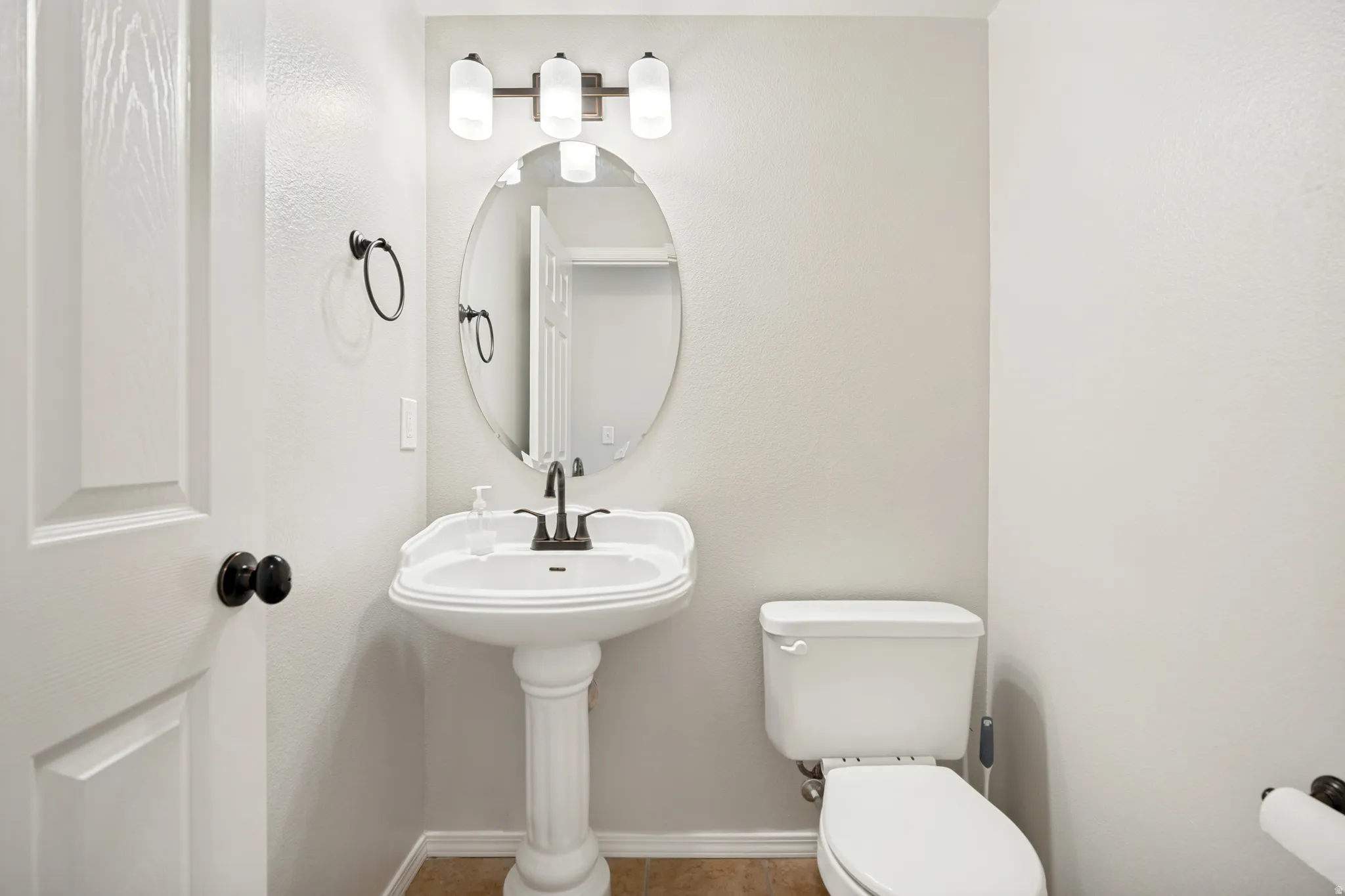 Powder room featuring a white pedestal sink with a dark bronze faucet, an oval mirror, and a three-light vanity fixture