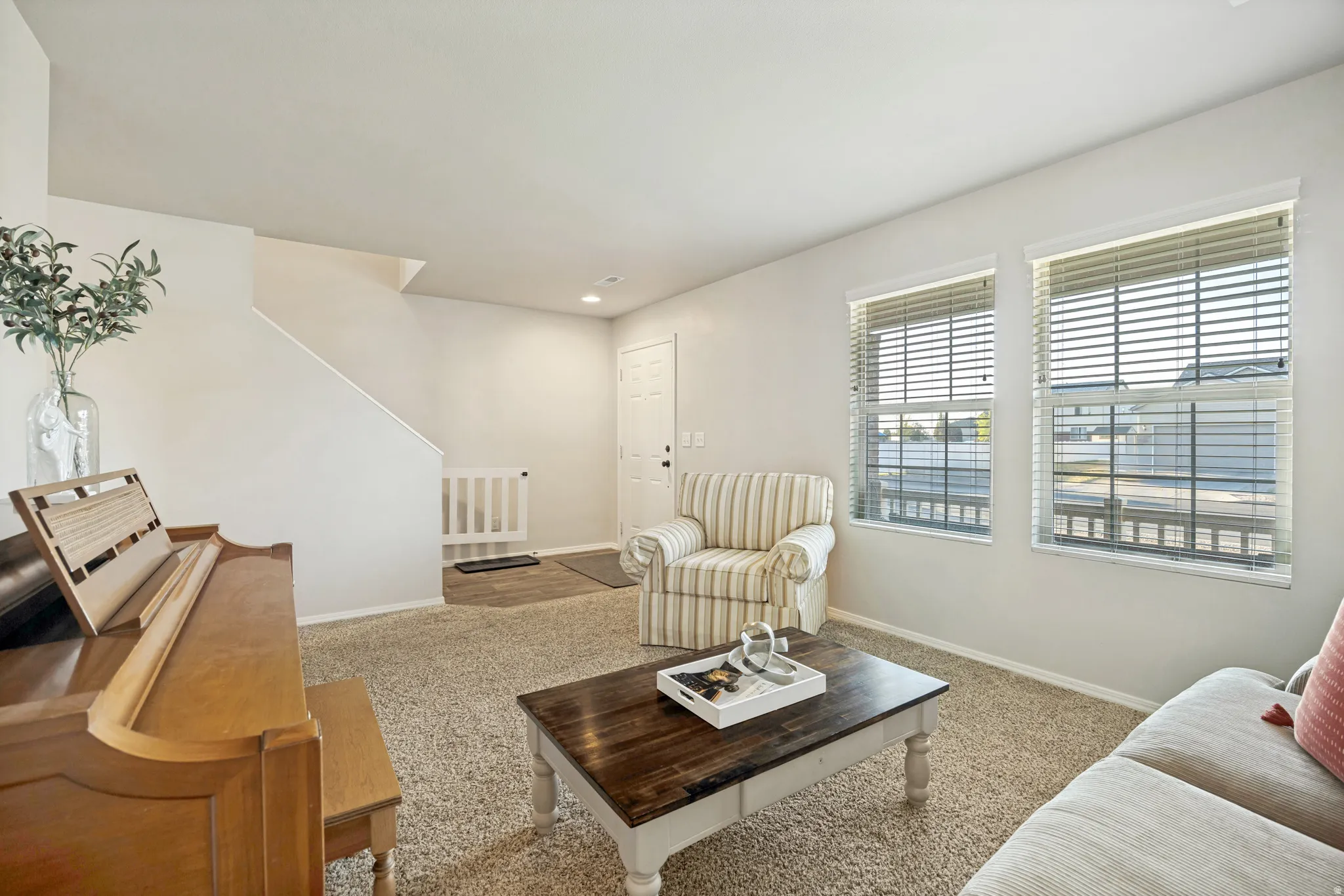 Carpeted living area featuring two windows with blinds, a white door, and a staircase