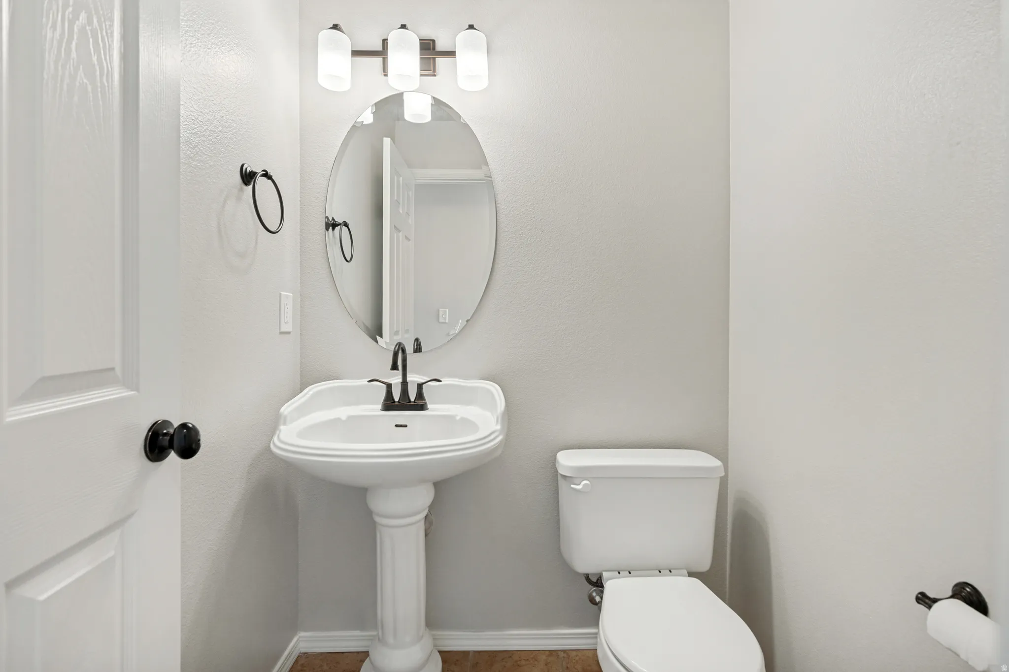 Pedestal sink with dark-toned fixtures, oval mirror, and a three-light vanity fixture
