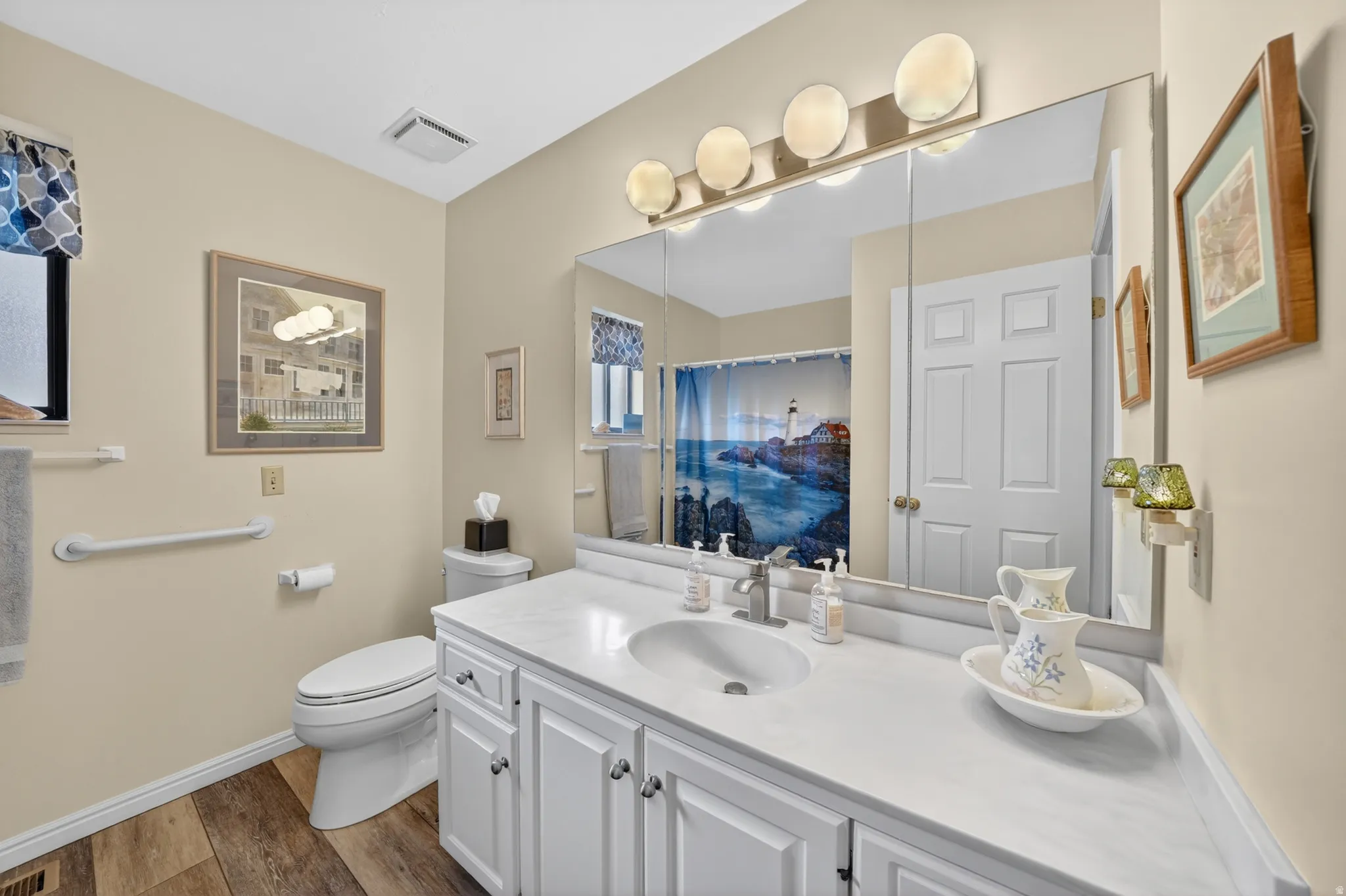 Bathroom featuring a white vanity with an integrated sink, a large mirror with a four-light fixture, wood-finish flooring, a toilet, and a window with a valance