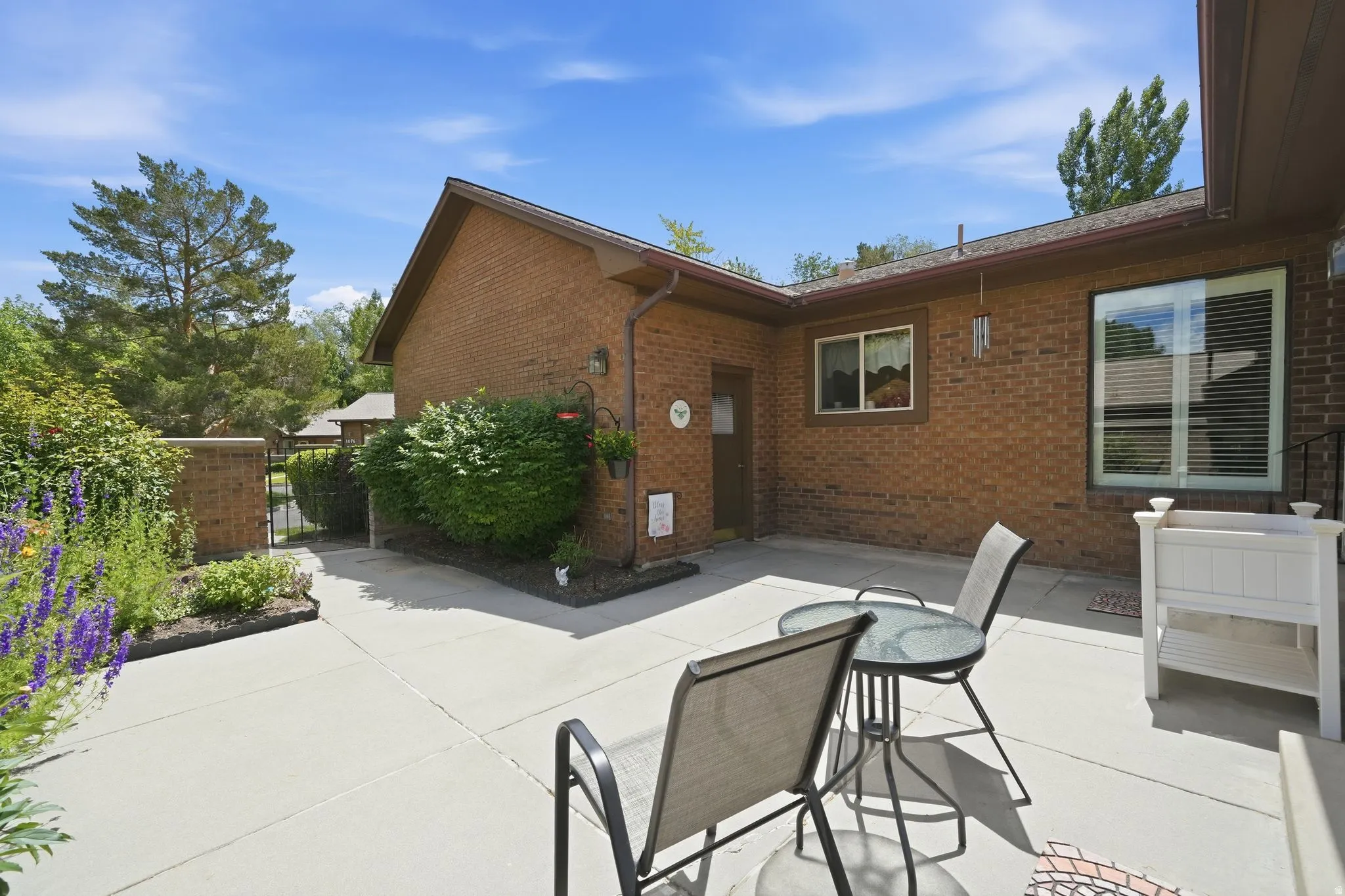 Private brick patio space featuring a concrete surface, surrounding garden beds with flowering plants and established shrubs, and a partial brick privacy wall with a metal gate