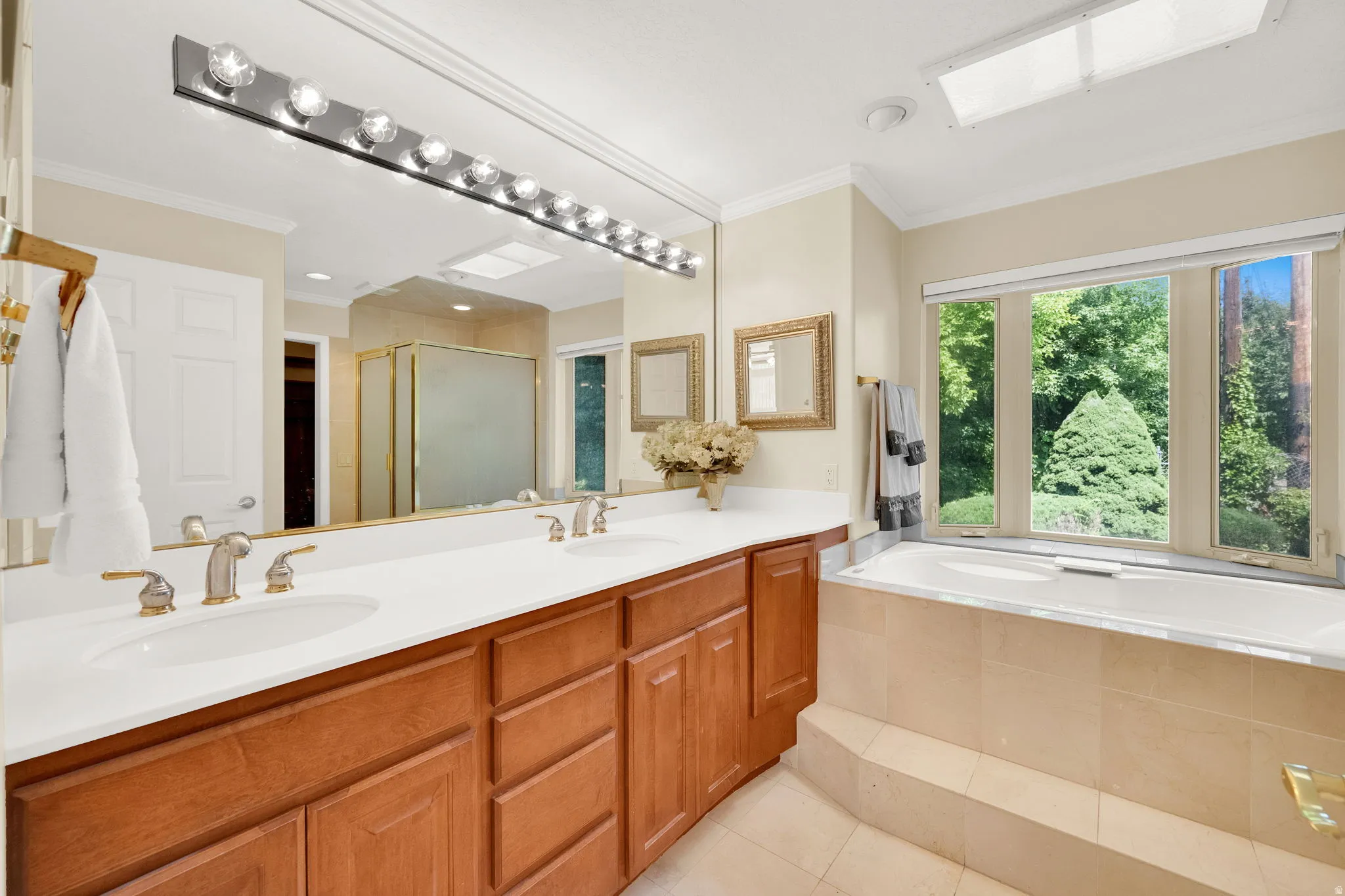 Spacious bathroom featuring a double vanity with wood cabinetry, a built-in soaking tub with tiled surround, a large mirror, and a skylight