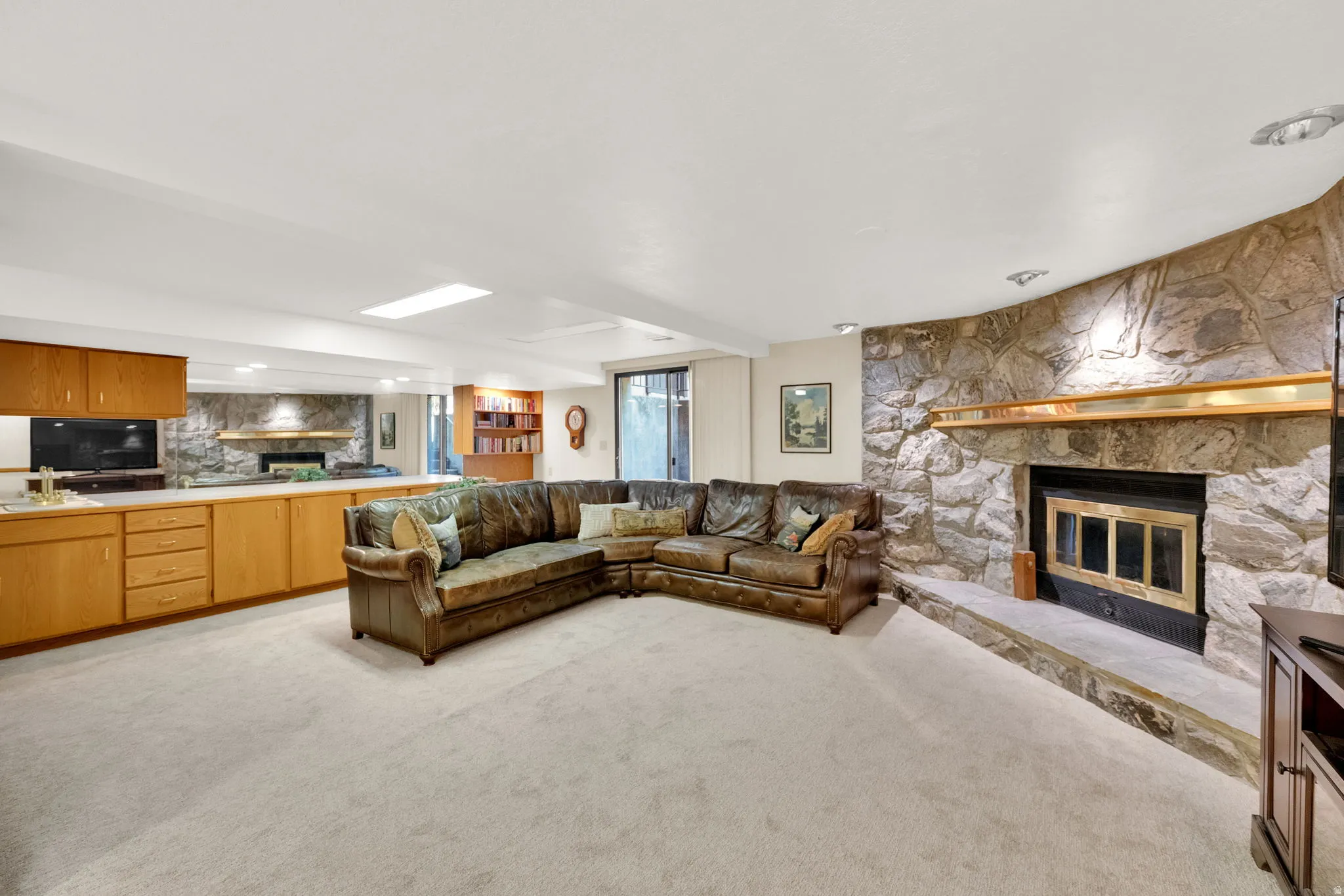 Spacious lower level featuring a stone-clad fireplace with a brass mantel, light-colored carpeting, built-in wooden cabinetry with a countertop, and recessed lighting