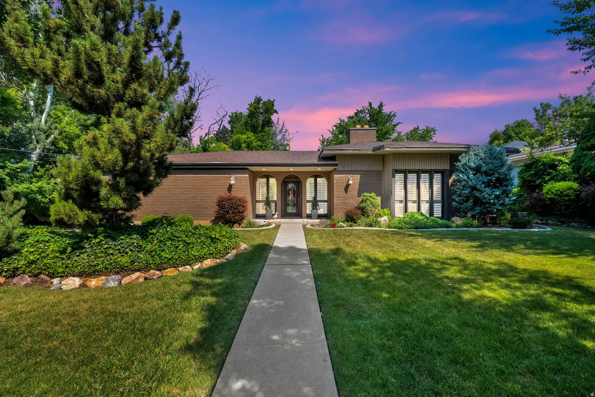 Front exterior featuring brick and panel siding, a concrete walkway, established landscaping, and mature trees