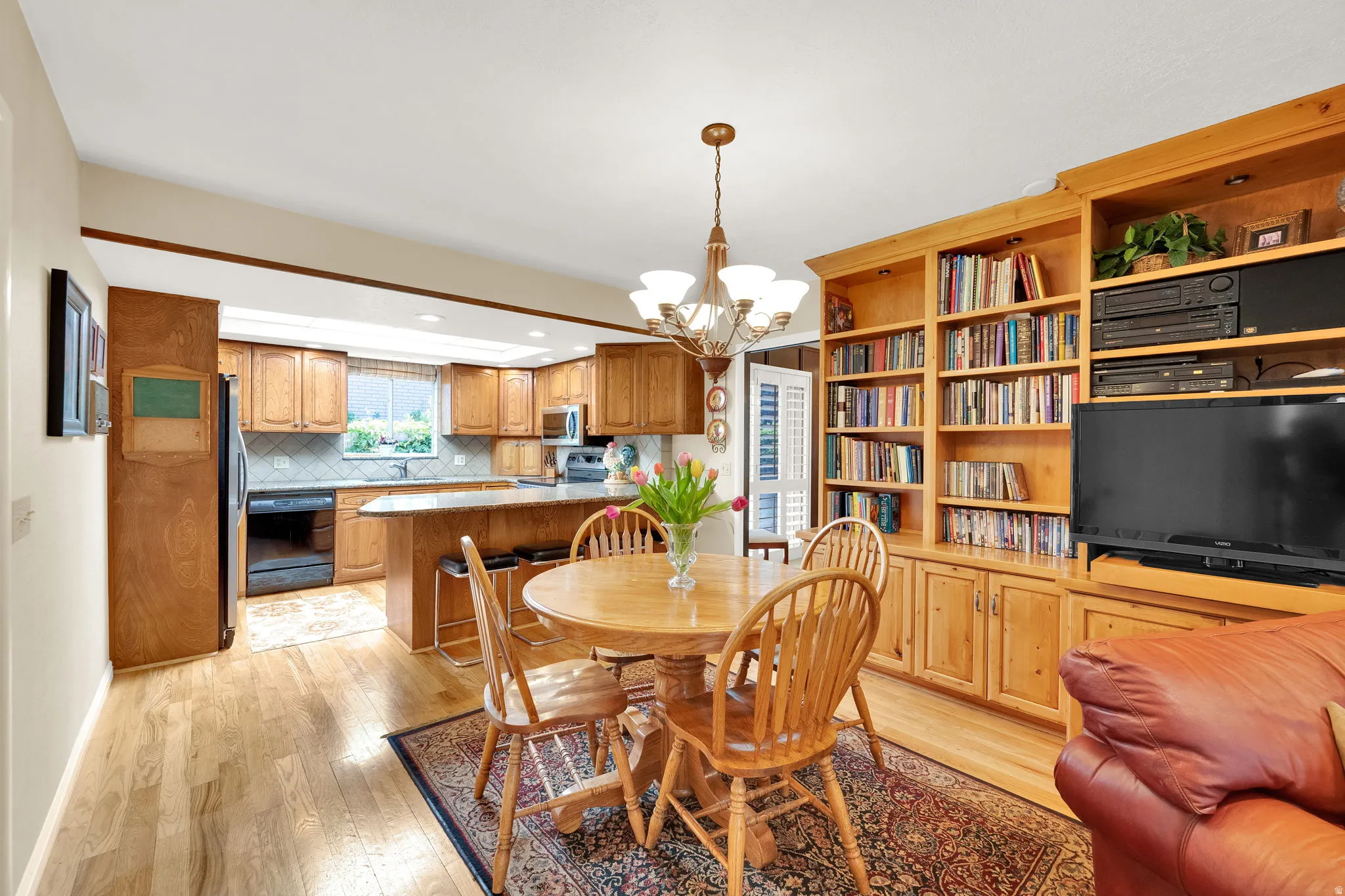 Open-concept living area featuring wood-finish flooring, a built-in entertainment center with integrated shelving, and a kitchen with wood cabinetry and a peninsula