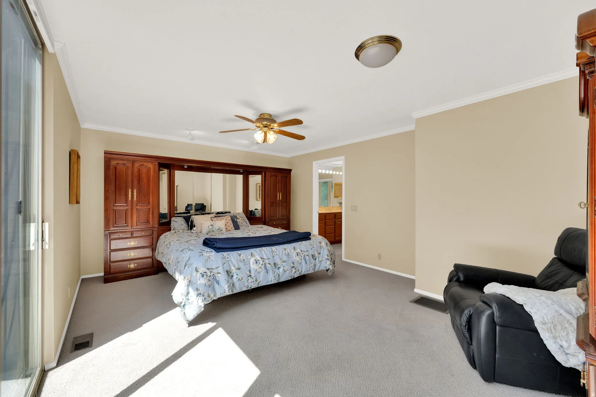 Spacious bedroom featuring neutral carpeting, crown molding, a ceiling fan with integrated lighting, and a built-in wood-finish storage unit with mirrored accents