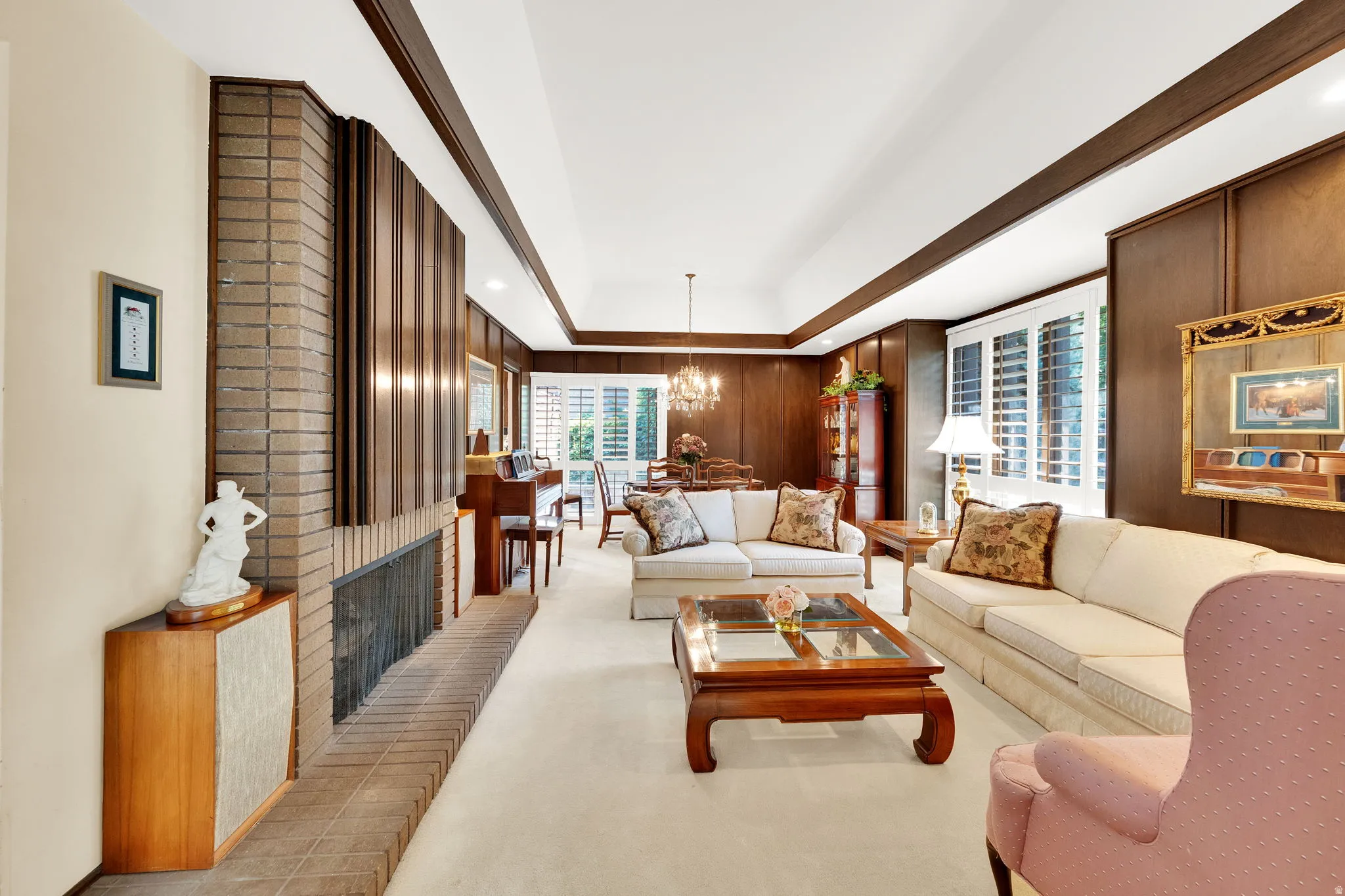 Spacious living area featuring a brick fireplace surround with wood paneling, dark wood accent walls, white plantation shutters, and a coffered ceiling
