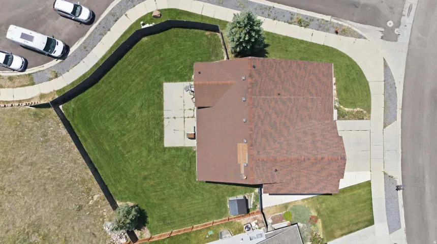Aerial view showcasing a brown-shingled roof, a large, manicured green lawn, concrete patio, and a paved driveway leading to the street