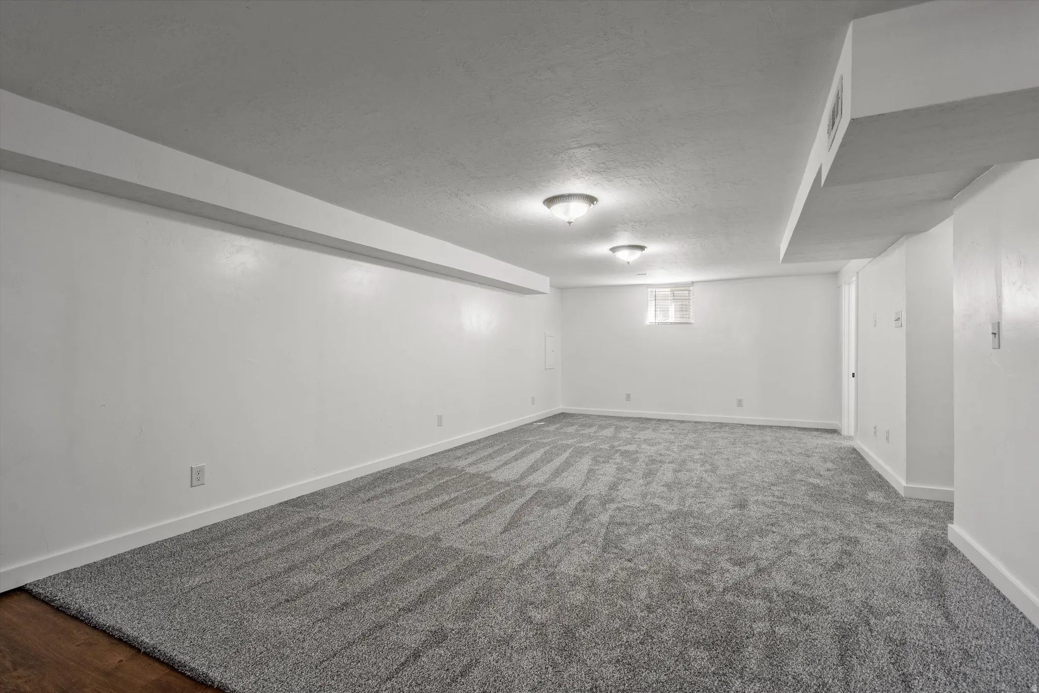Spacious carpeted room with neutral white walls, ceiling-mounted lighting fixtures, and a high window providing natural light