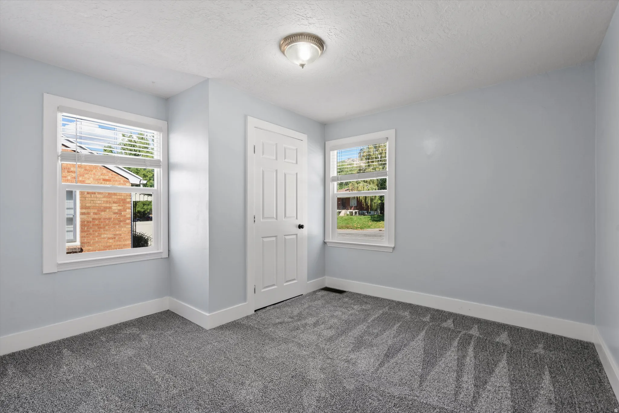 Light gray walls complement the textured gray carpet in this room featuring two windows with white blinds, a white door with black hardware, and a ceiling-mounted light fixture