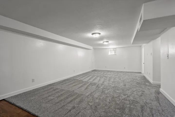 Spacious carpeted room with neutral white walls, ceiling-mounted lighting fixtures, and a high window providing natural light