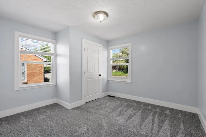 Light gray walls complement the textured gray carpet in this room featuring two windows with white blinds, a white door with black hardware, and a ceiling-mounted light fixture