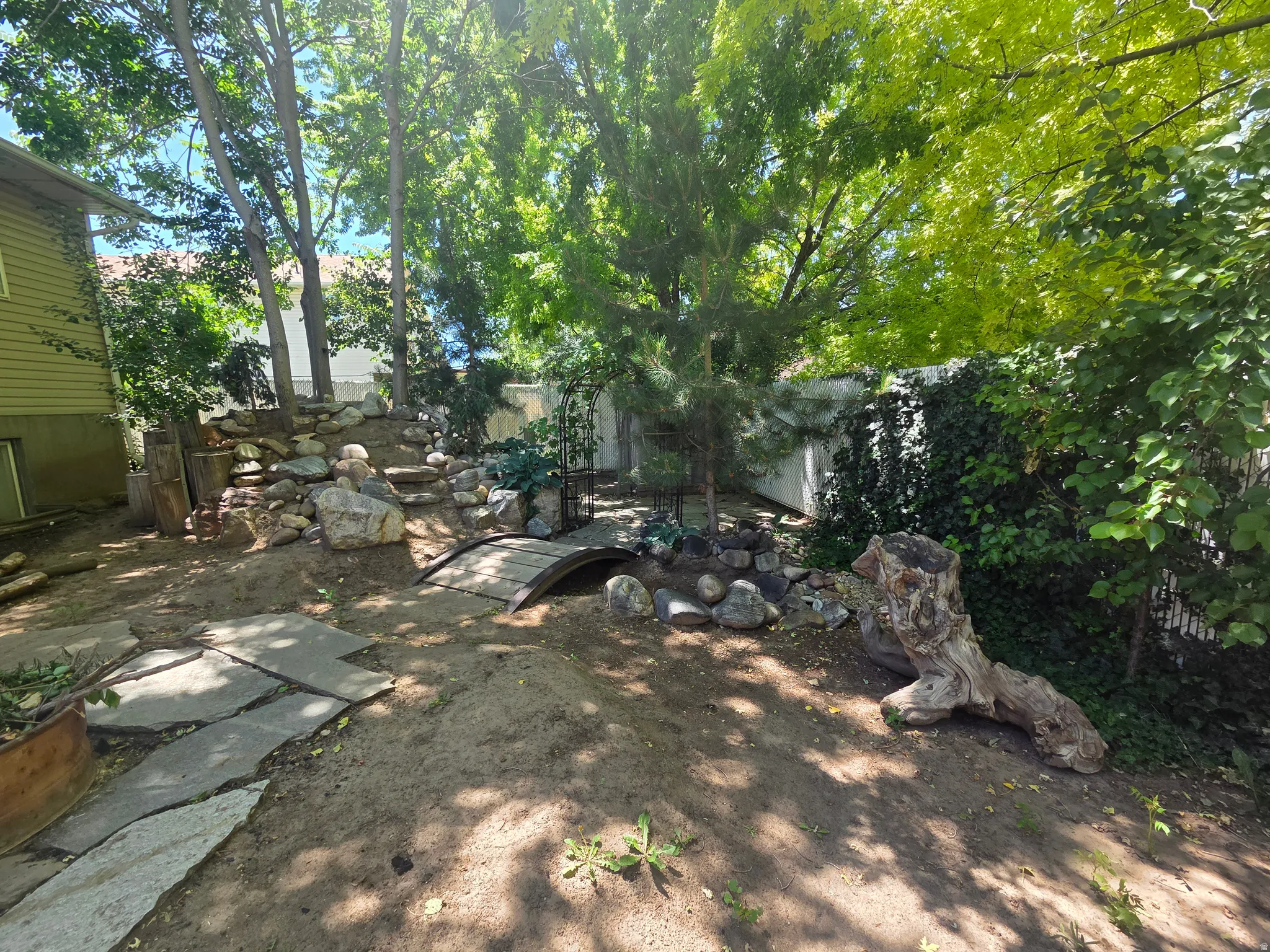 Terraced backyard featuring stone hardscaping, a decorative garden bridge, and mature trees providing natural shade