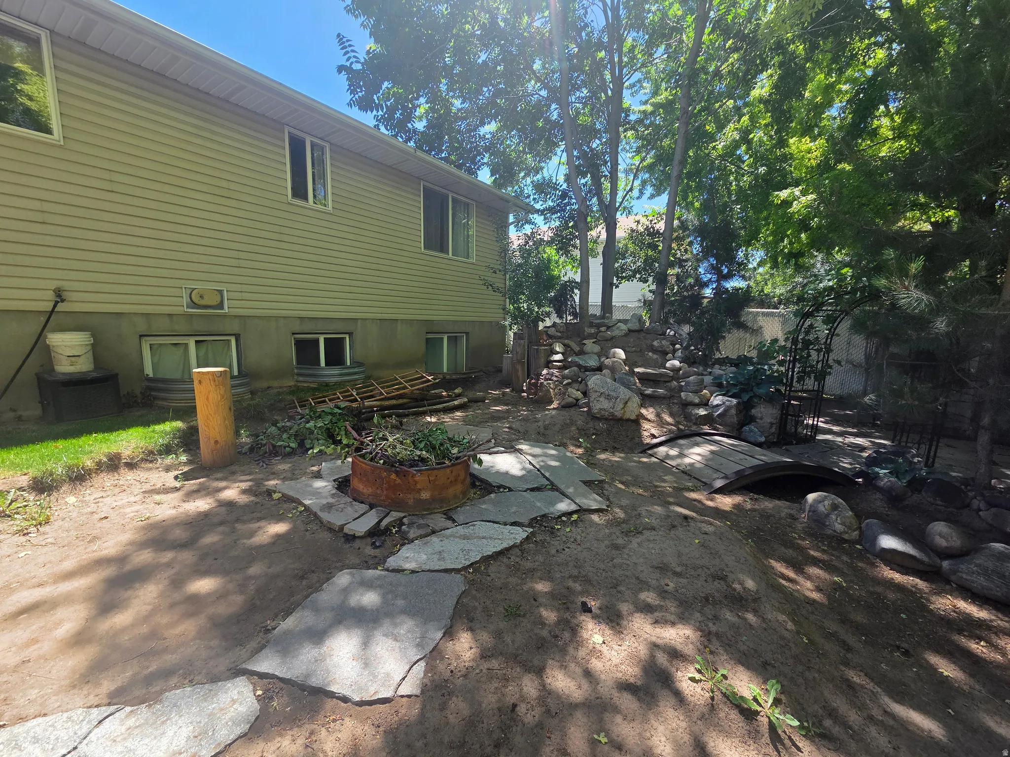 Shaded backyard featuring mature trees, a stone pathway, a decorative wooden bridge, and a rock garden