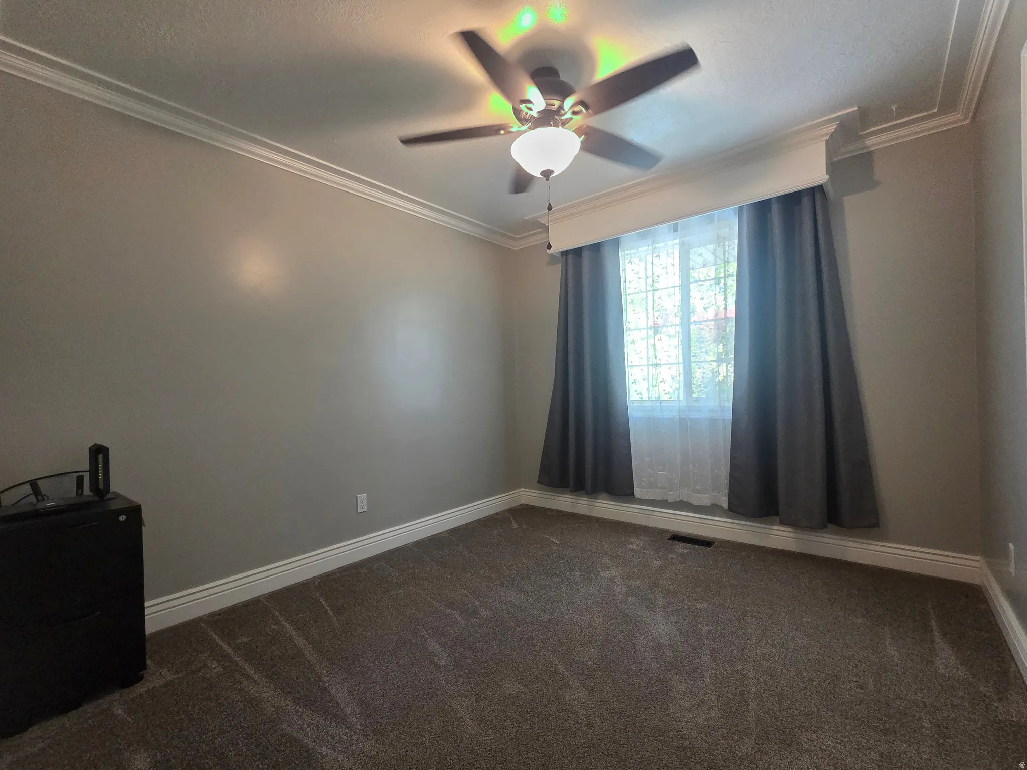 Room featuring crown molding, neutral wall color, textured carpet, a window with window treatments, and a ceiling fan with integrated lighting