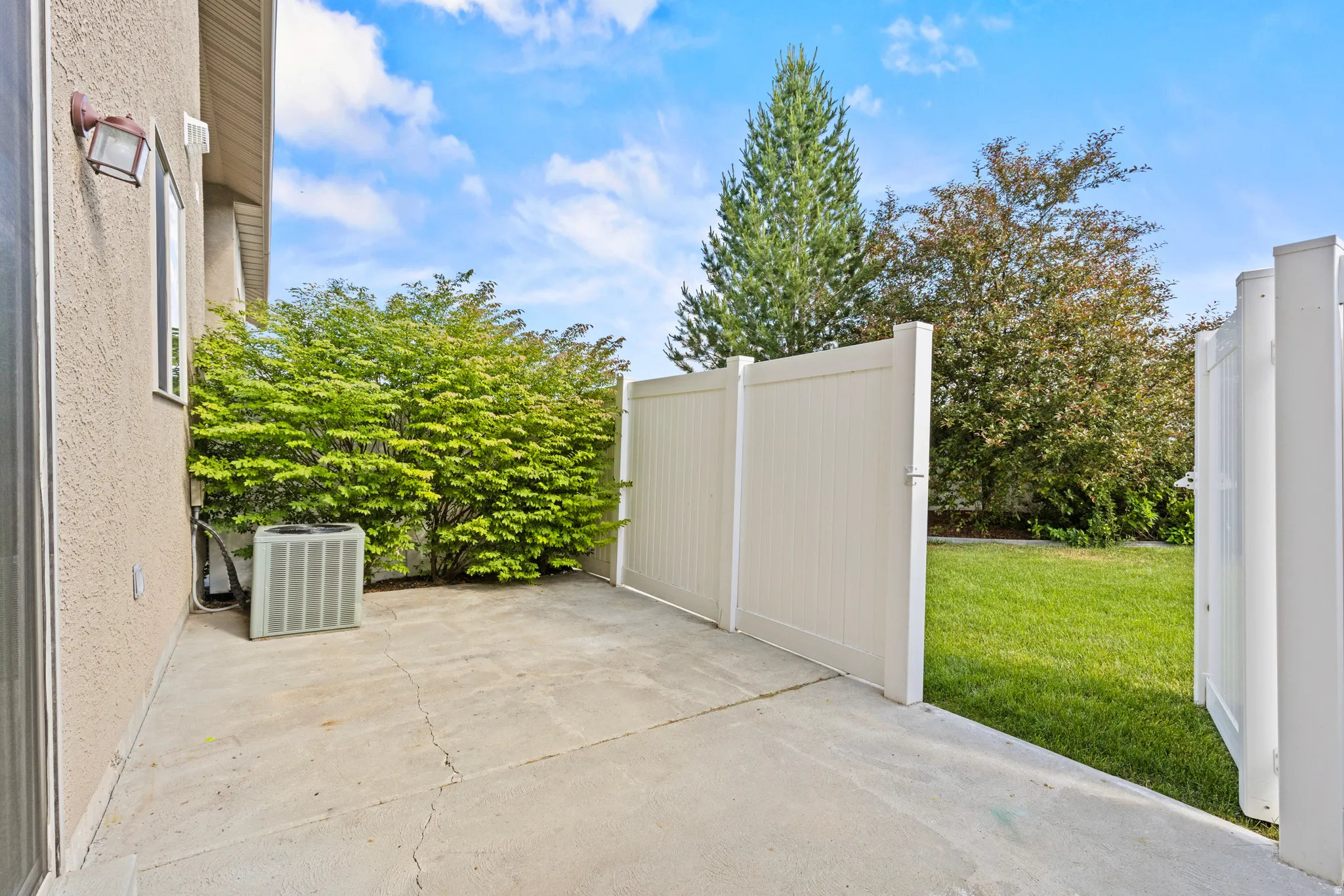 Concrete patio area featuring a textured exterior wall, an exterior light fixture, and surrounding mature landscaping