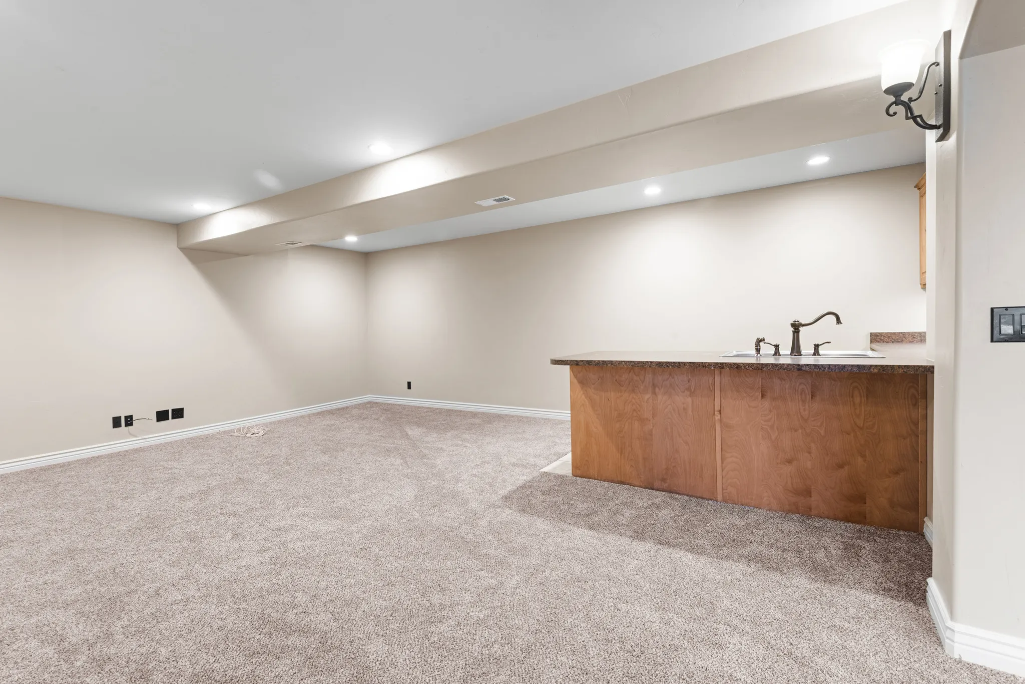 Finished basement room featuring neutral carpeting, recessed lighting, and a built-in wet bar with wood-finish cabinetry, a stone-look countertop, and a single-basin sink with a dark bronze faucet