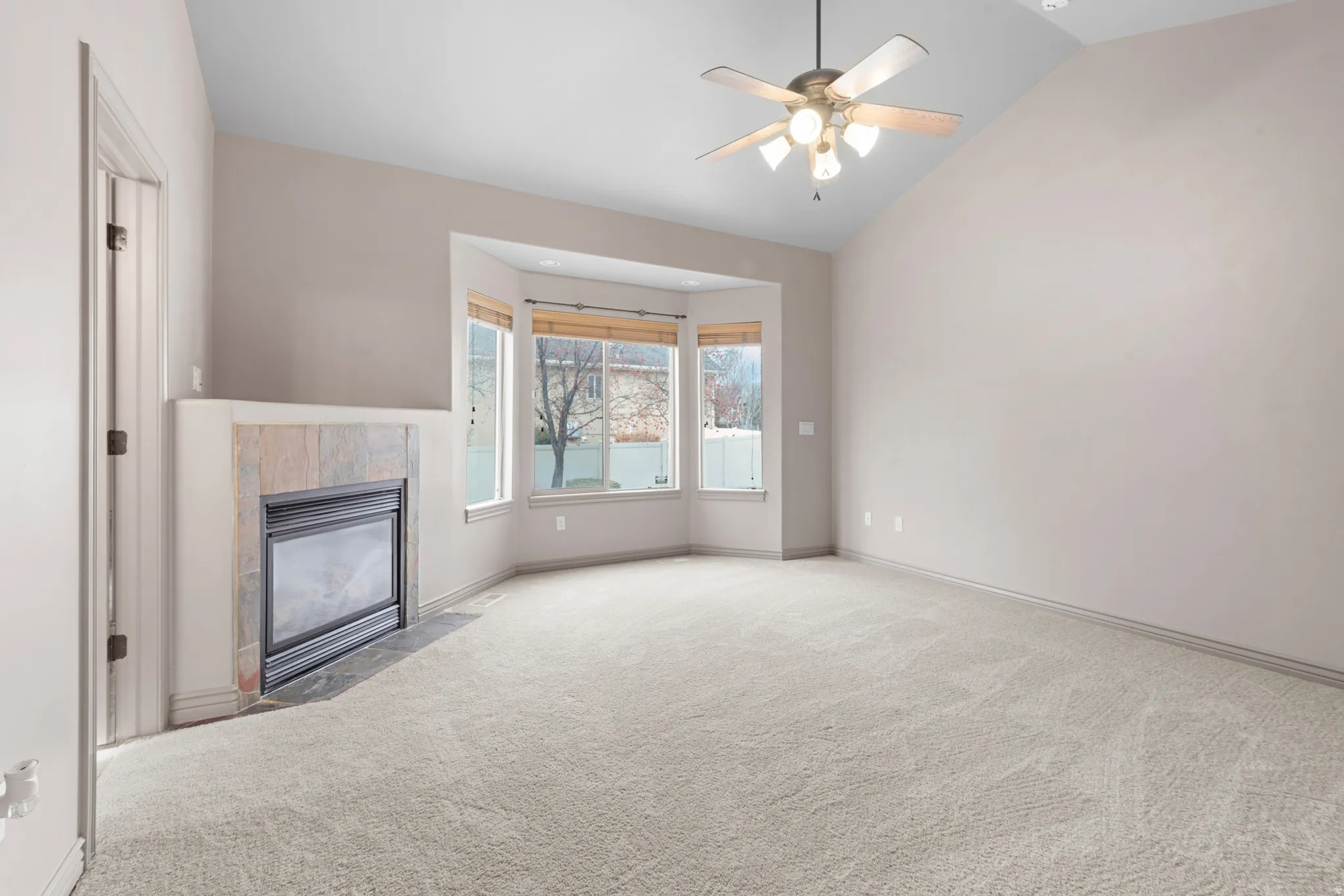Carpeted room featuring a slate-tiled fireplace, a bay window with blinds, a vaulted ceiling, and a ceiling fan with integrated lighting
