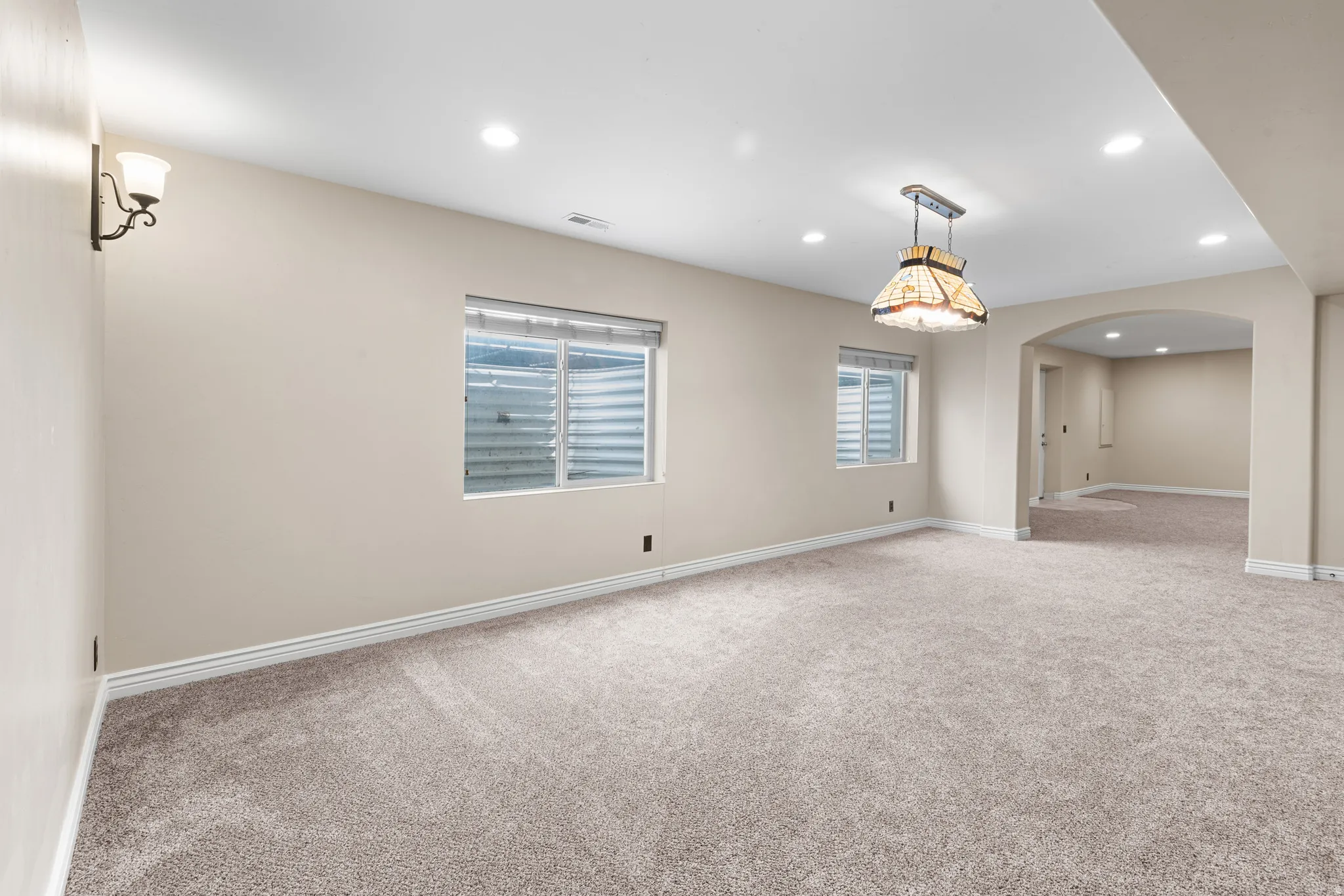 Spacious room featuring carpet flooring, recessed lighting, two windows with blinds, a decorative ceiling fixture, and a wall sconce