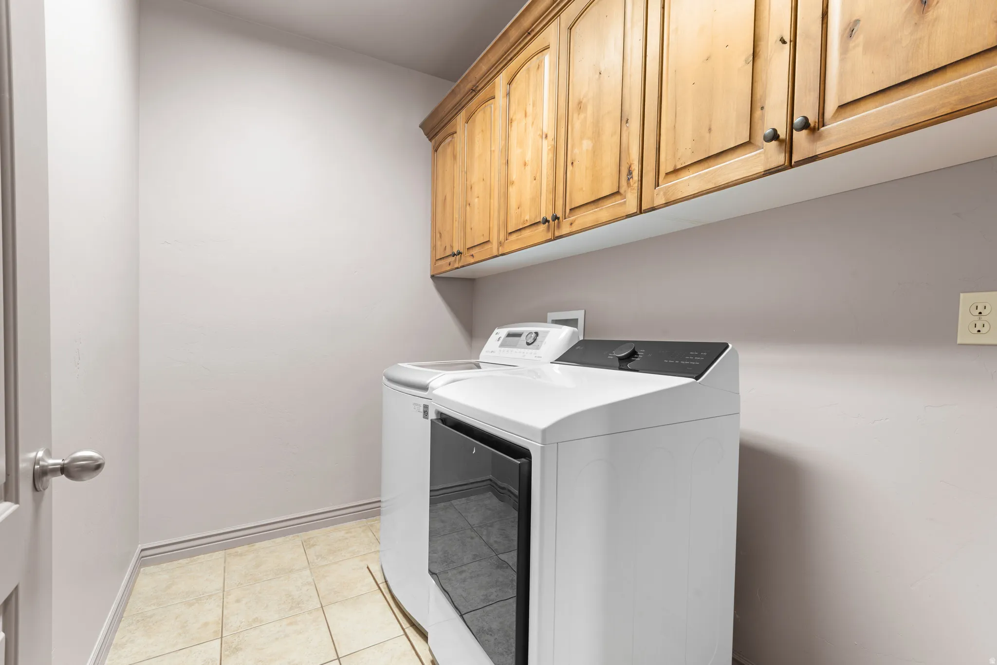 Laundry area featuring wood-finish overhead cabinetry with dark hardware, neutral wall paint, and light-toned tile flooring