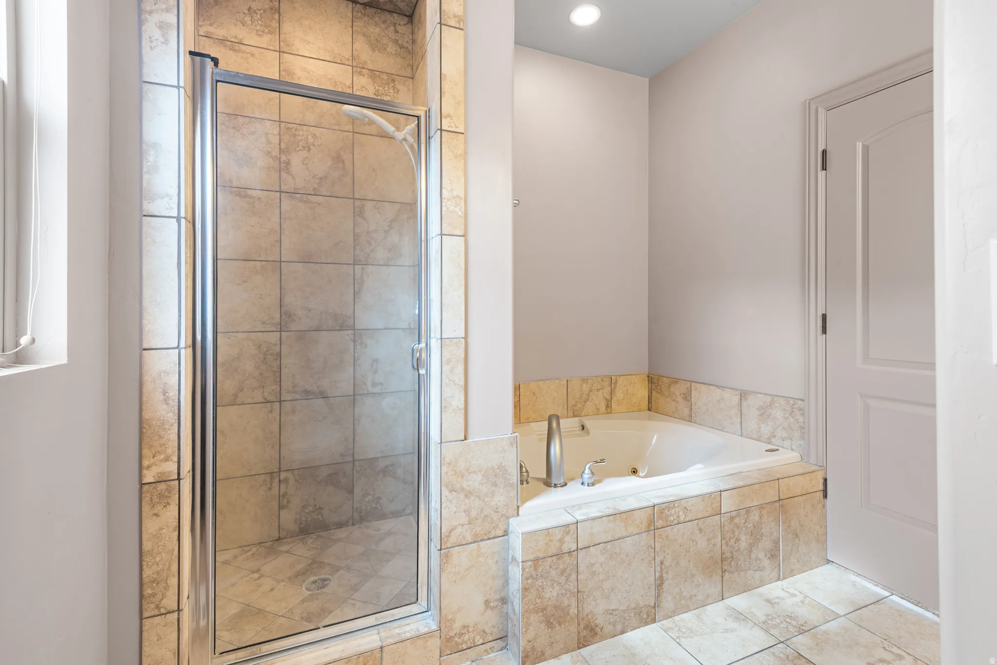 Spacious bathroom featuring a built-in corner soaking tub with tiled surround, a separate tiled shower enclosure with a glass door, and coordinated neutral tile flooring
