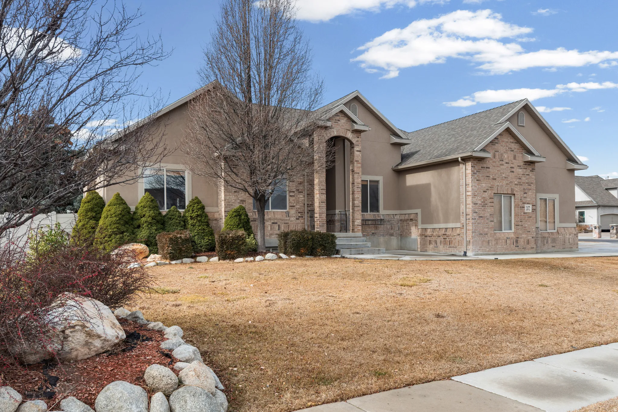 Stucco and brick facade residence featuring a prominent arched entryway, gabled rooflines, and a landscaped front yard with evergreen shrubs and rock accents