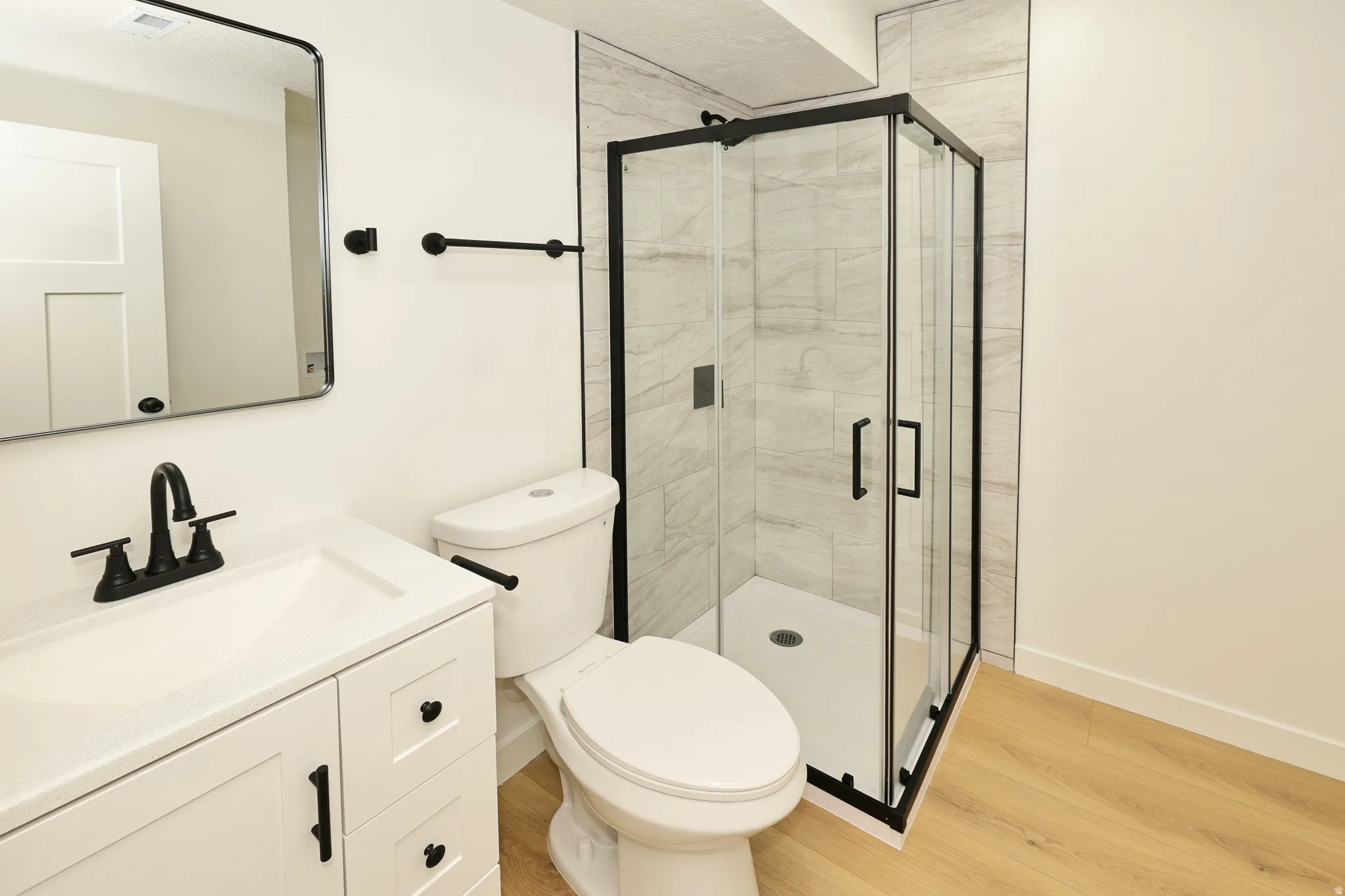 Modern bathroom featuring a corner glass-enclosed shower with tile surround, a white vanity with integrated sink, matte black fixtures, and wood-finish flooring