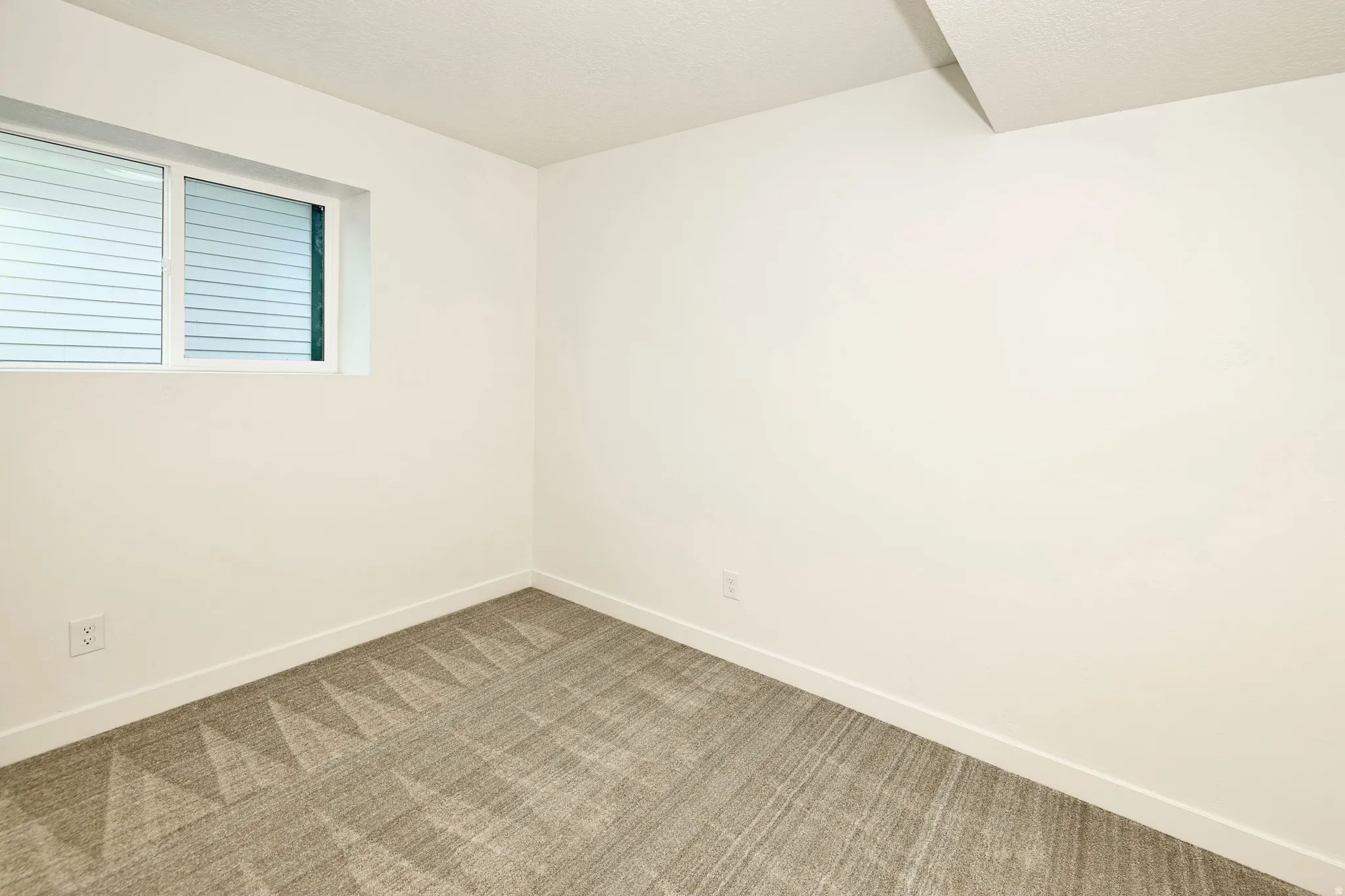 Neutral-toned room featuring a single window with blinds, light-colored walls, and a textured gray carpet