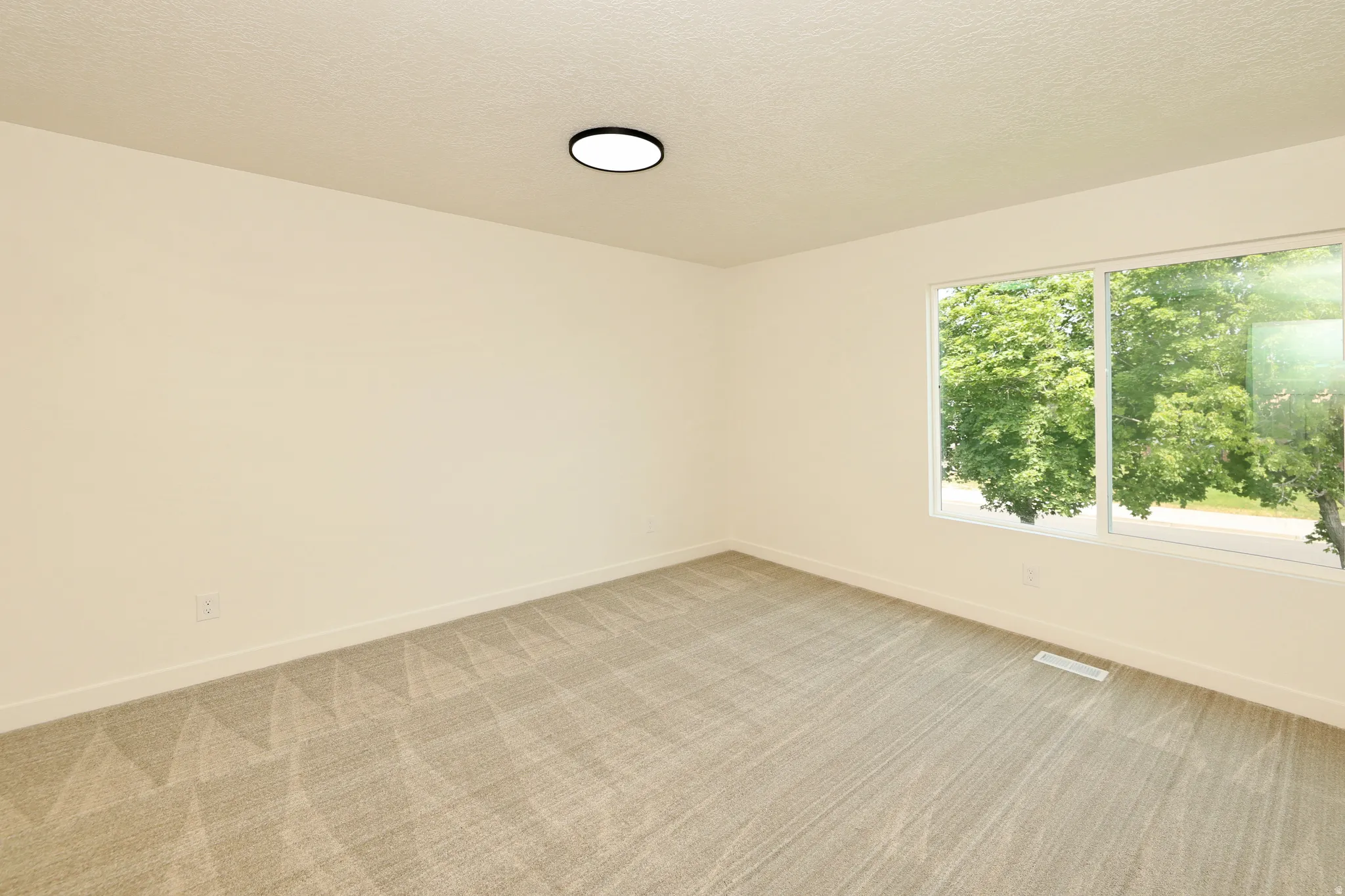 Bright room featuring a large window with green tree scenery, light-colored walls, and gray patterned carpeting
