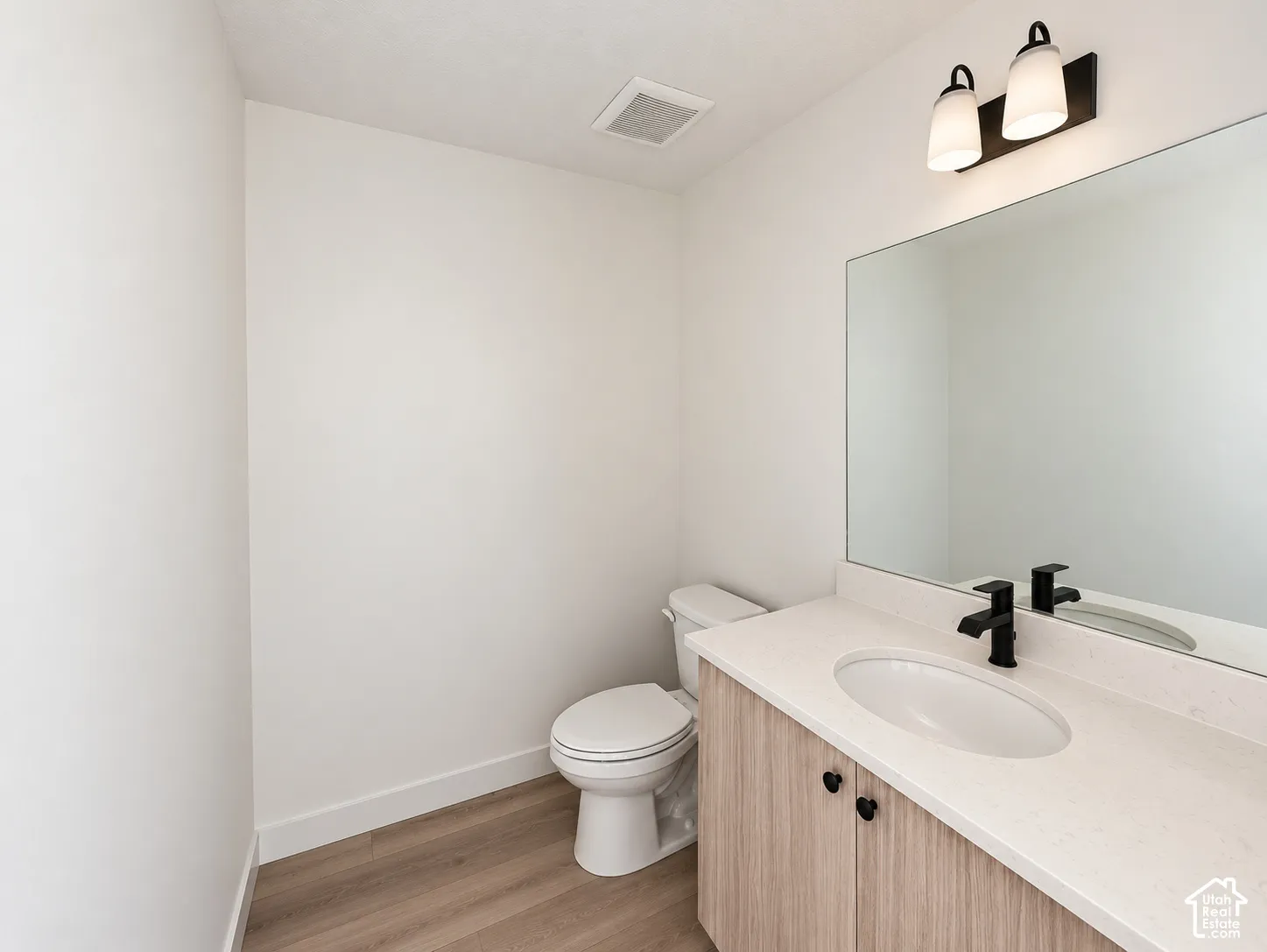 Wood-finish flooring, light wood-look vanity with black hardware, white countertop, black faucet, and rectangular mirror