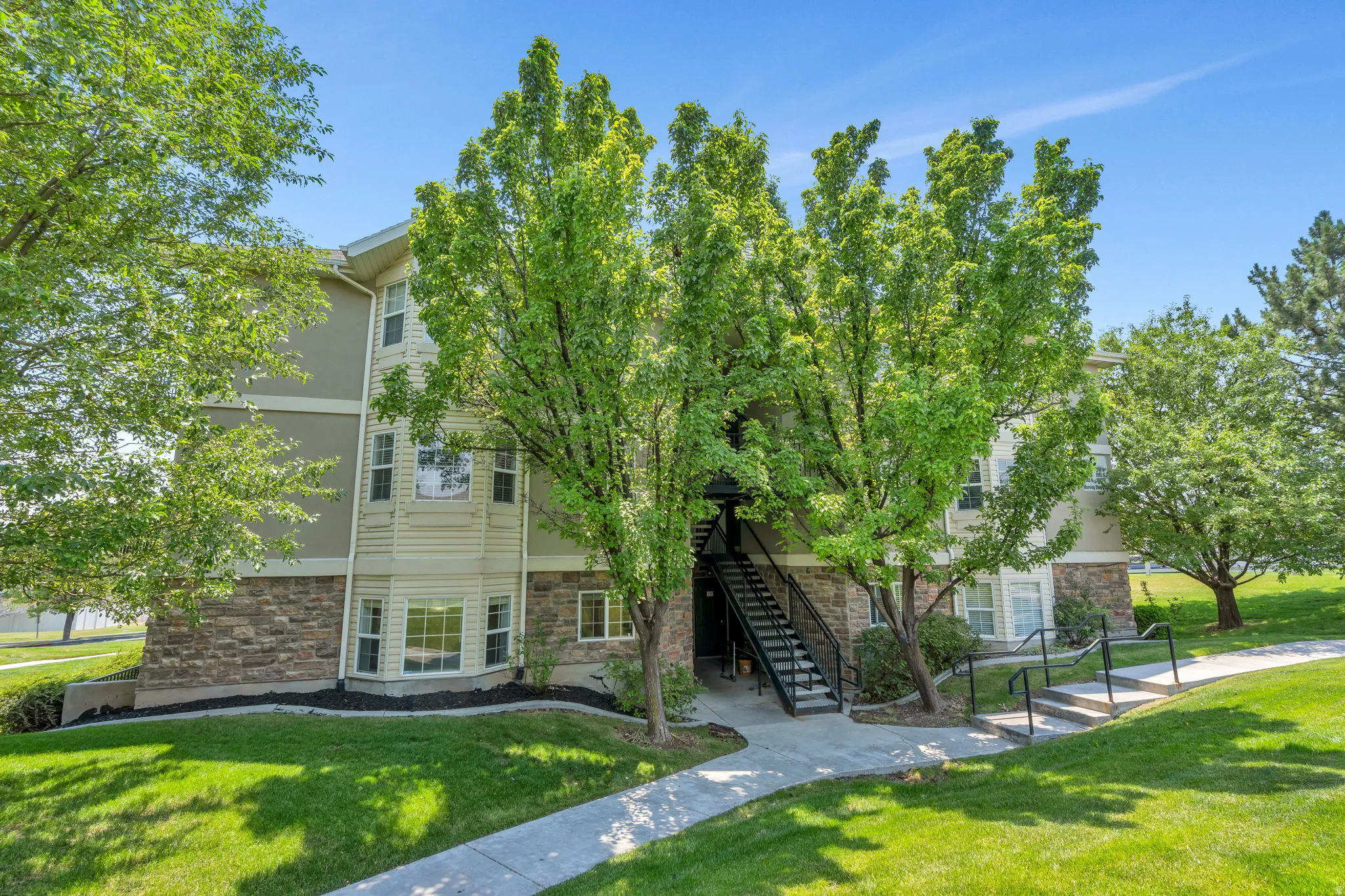 Multi-story residential building featuring stone veneer and siding exterior, established green landscaping, an exterior stairway with metal railings, and a concrete pathway