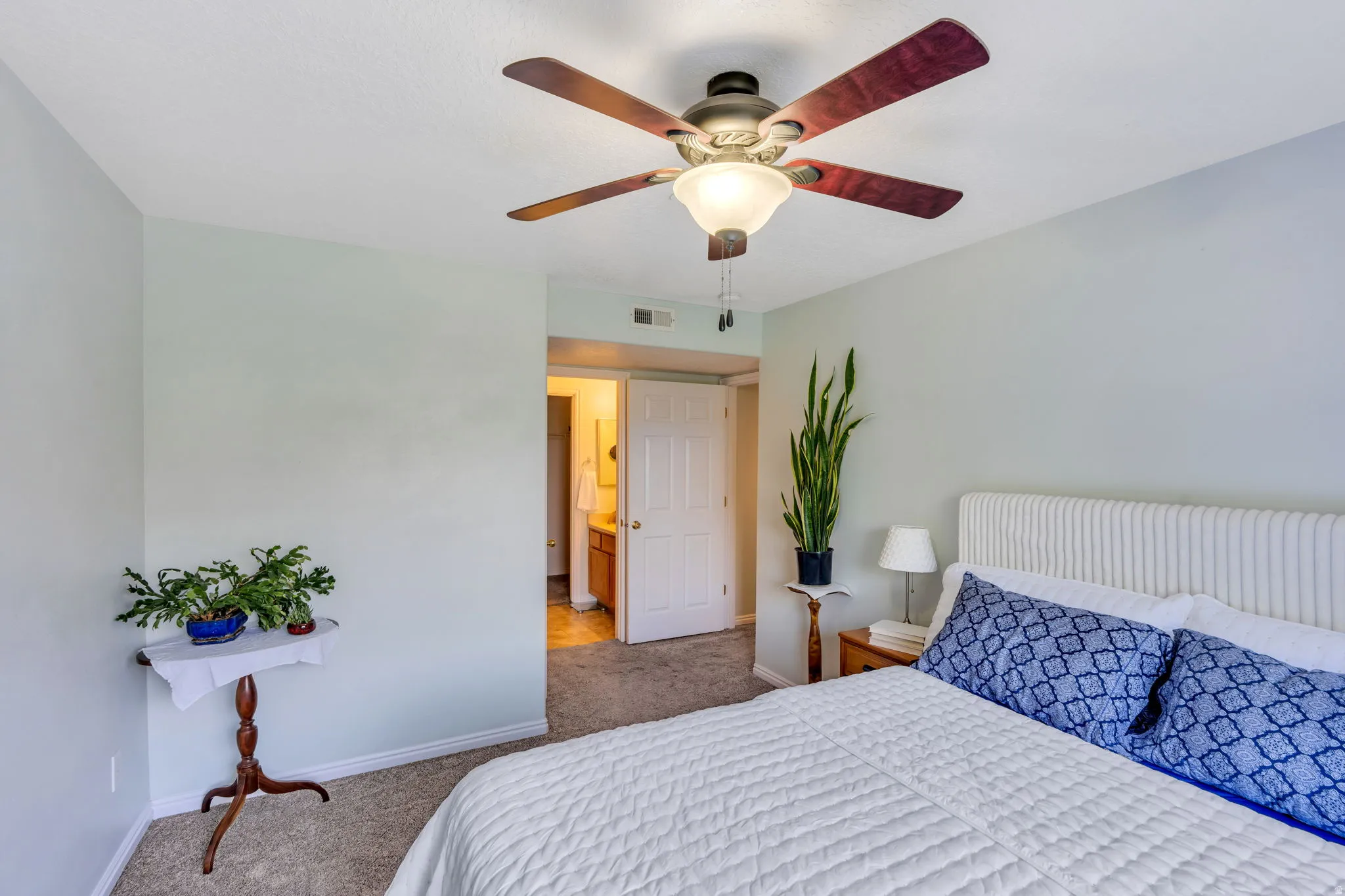 Carpeted room featuring a ceiling fan with light fixture, light blue wall paint, white baseboards, and an open doorway leading to an adjacent space with wood-finish cabinetry